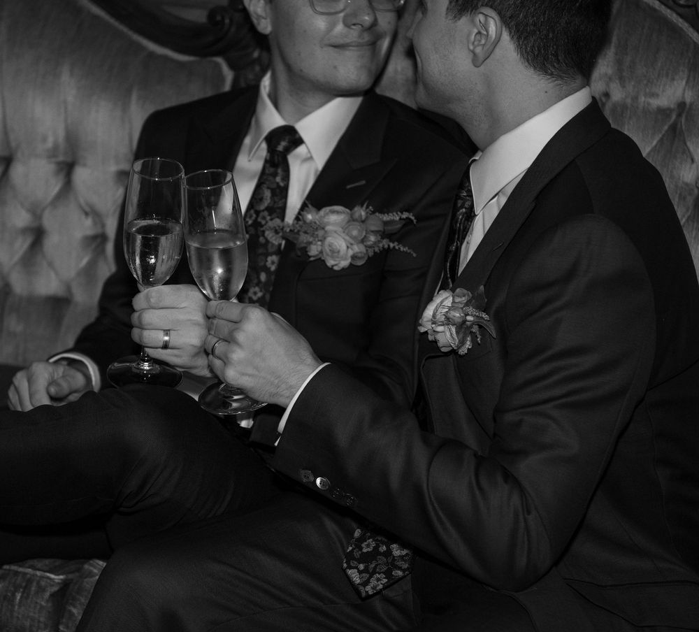 Two grooms in suits toasting with champagne glasses, wearing boutonnieres, in a black and white wedding photo.