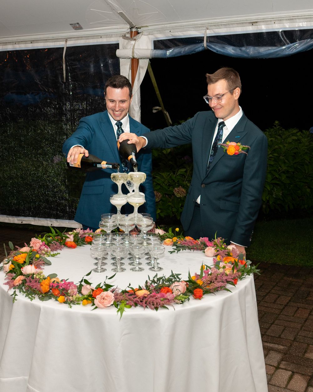Two grooms in teal suits pour champagne tower at wedding reception with floral arrangements.