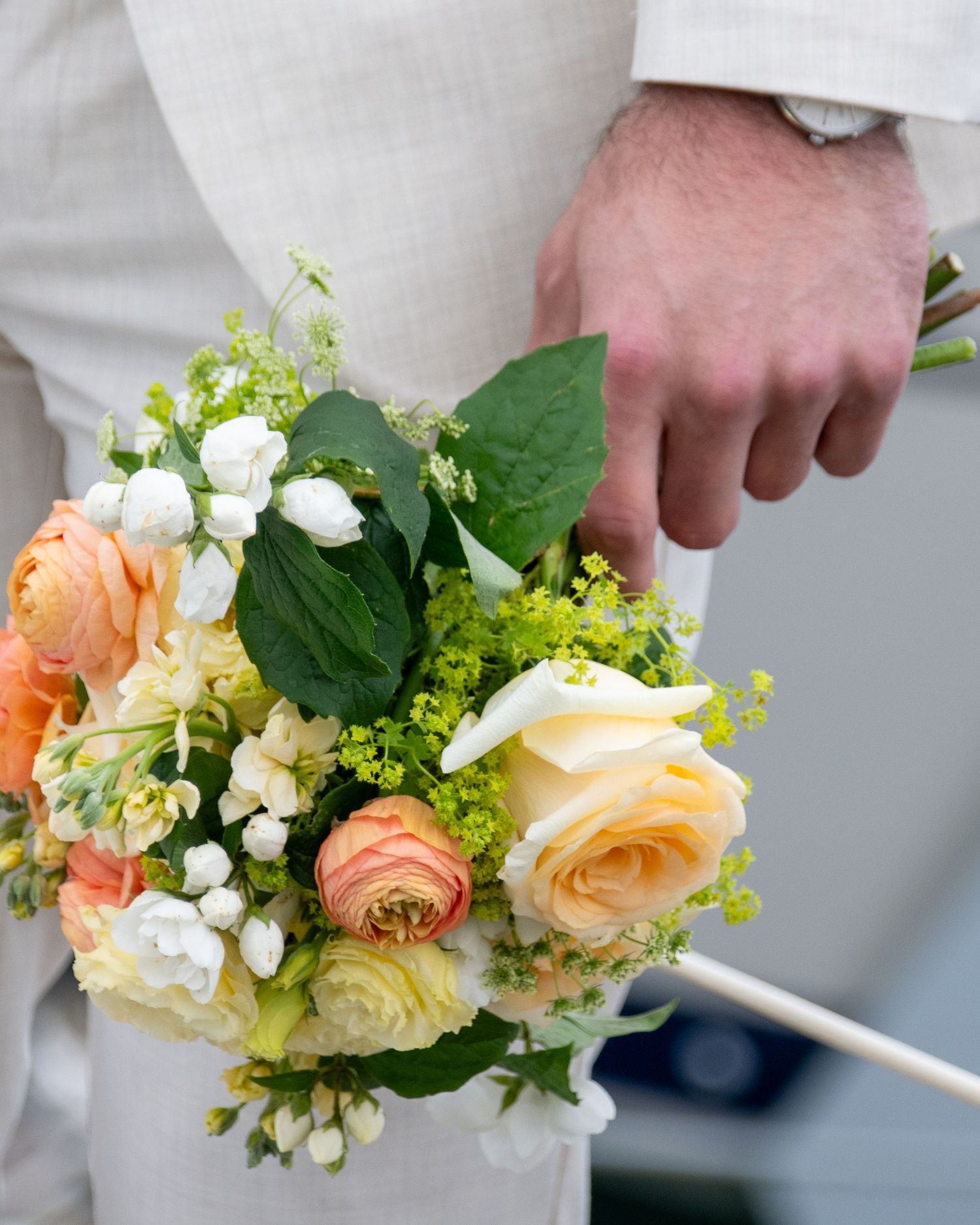 A hand holds an elegant wedding bouquet featuring peach roses, white flowers and green foliage against a light background.