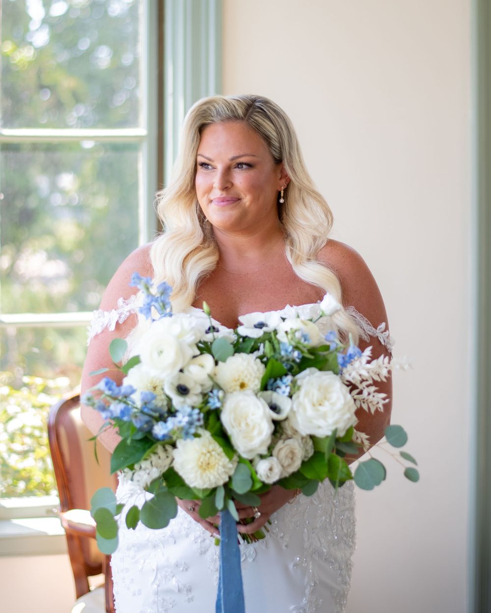 Bride in lace off-shoulder gown holding white and blue floral bouquet with eucalyptus, standing near a bright window.