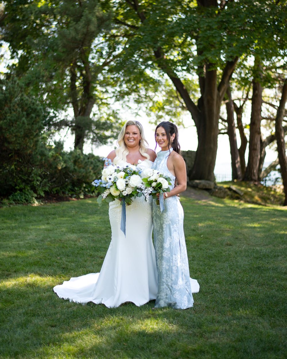 Two brides pose together on green lawn, one in fitted white gown holding white floral bouquet, other in lace dress, trees behind.