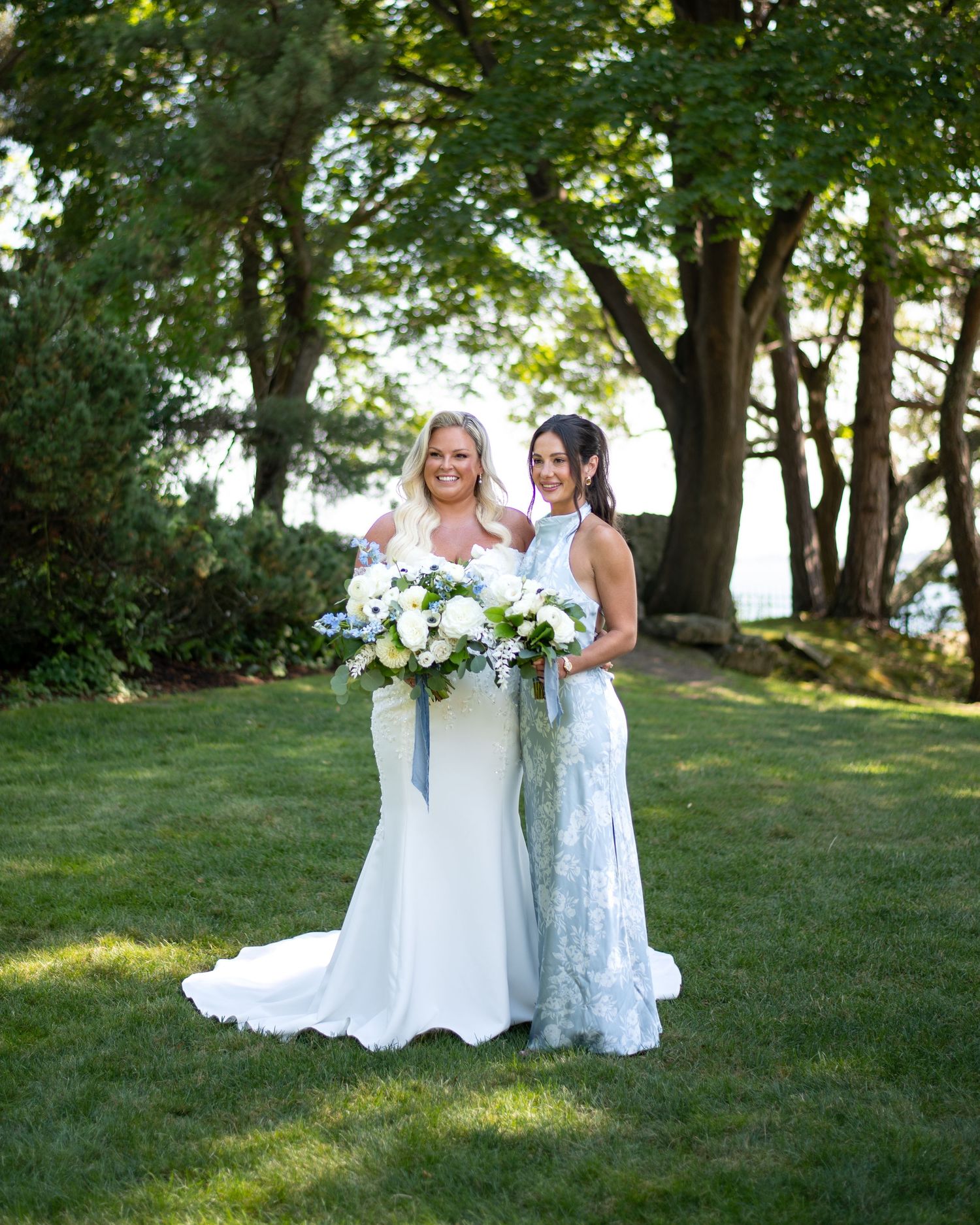 Two brides pose together on green lawn, one in fitted white gown holding white floral bouquet, other in lace dress, trees behind.