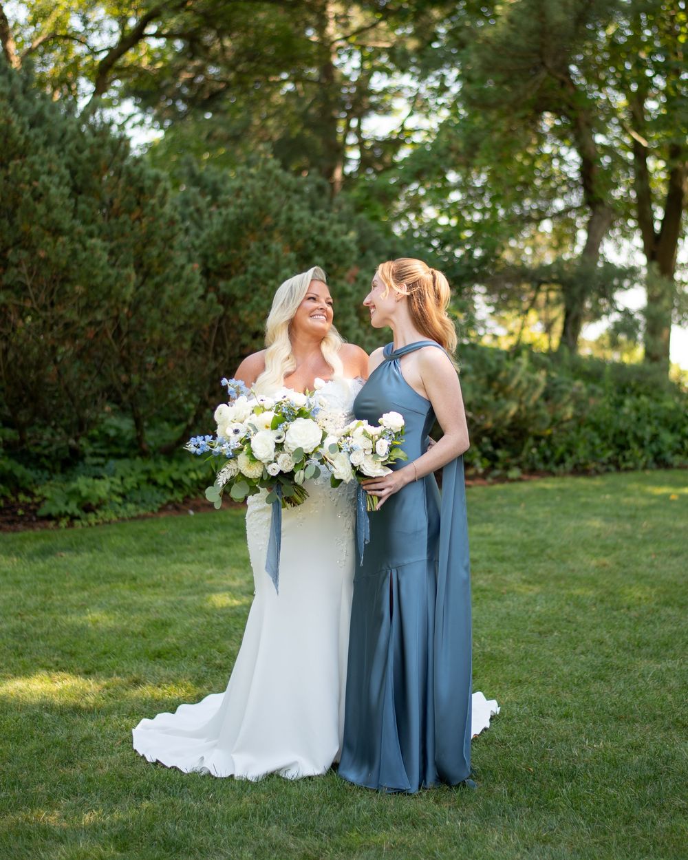 Bride in white gown and bridesmaid in dusty blue dress holding white floral bouquets smiling at each other outdoors.