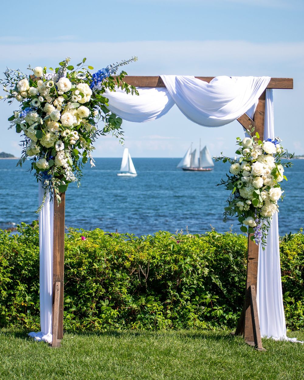 Wooden wedding arch adorned with white and blue flowers, draped white fabric, overlooking a serene ocean with sailboats.