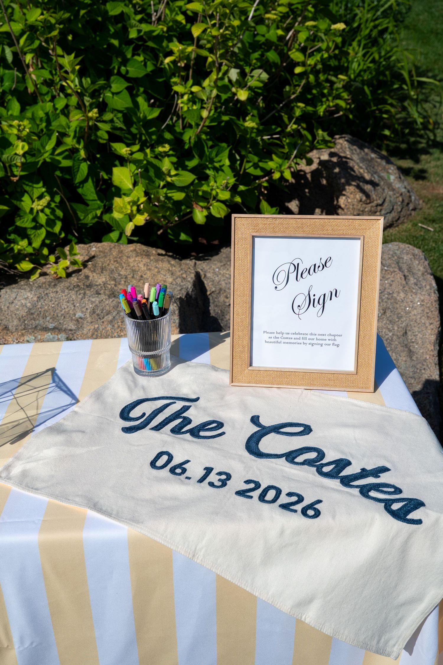 Wedding guest book table with "The Costes 06.13.2026" cloth, framed "Please Sign" sign, and colorful markers outdoors.