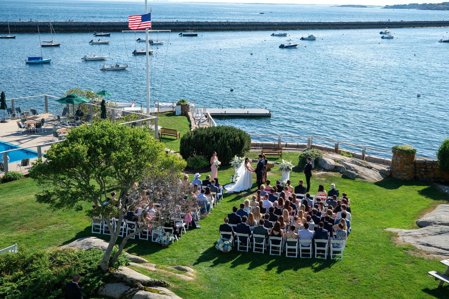 Outdoor waterfront wedding ceremony on a green lawn overlooking a harbor with boats and an American flag.