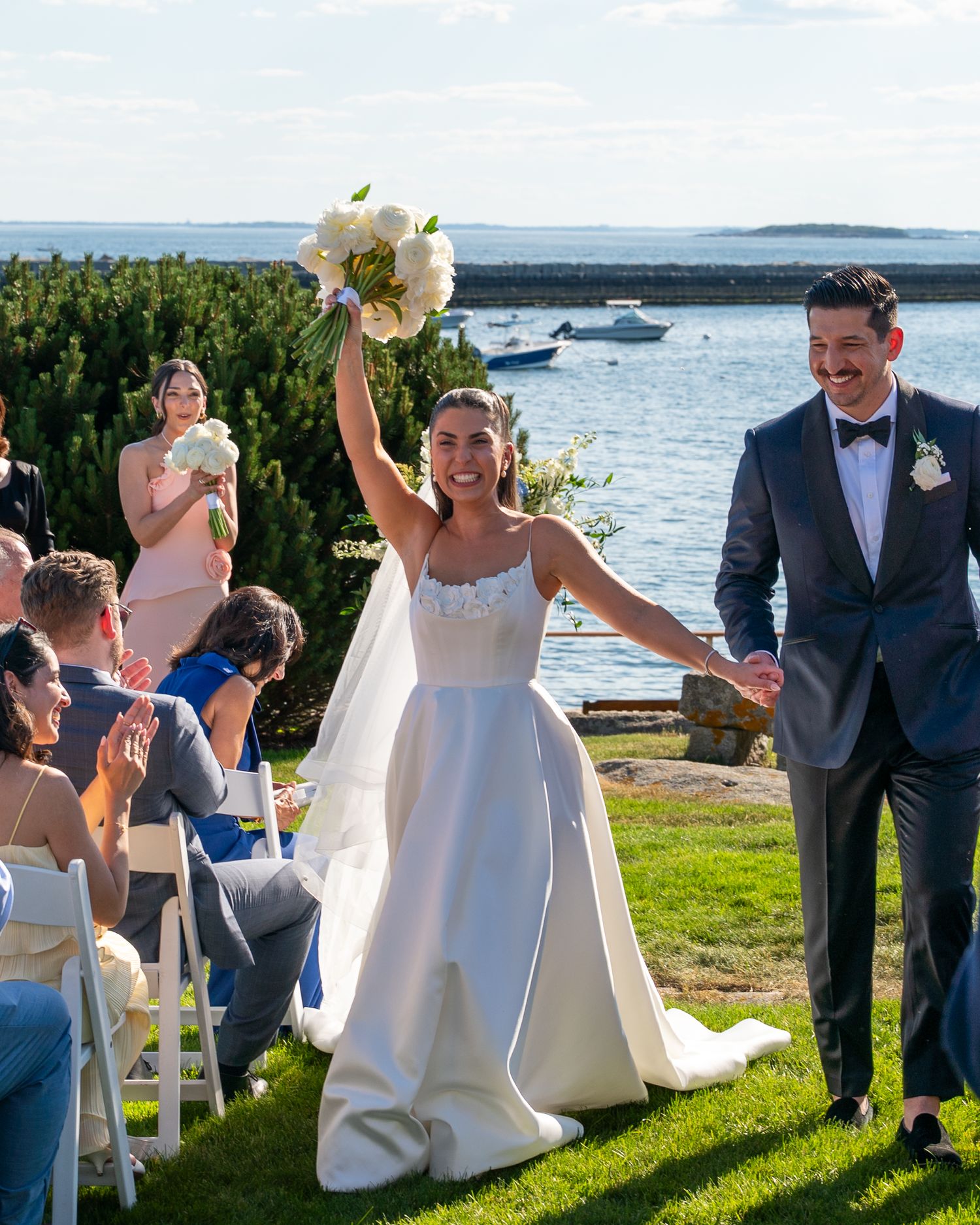Joyful bride raises bouquet while walking with groom after outdoor waterfront wedding ceremony on sunny day.