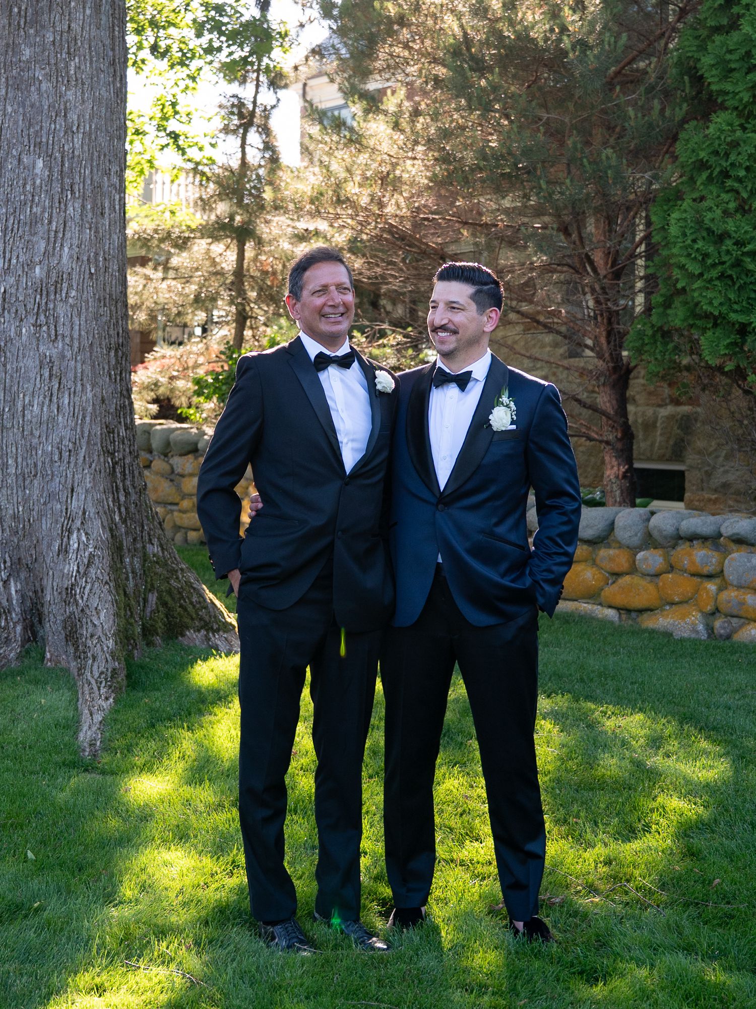 Two men in black tuxedos with bow ties smile together outdoors on green grass at a wedding celebration.