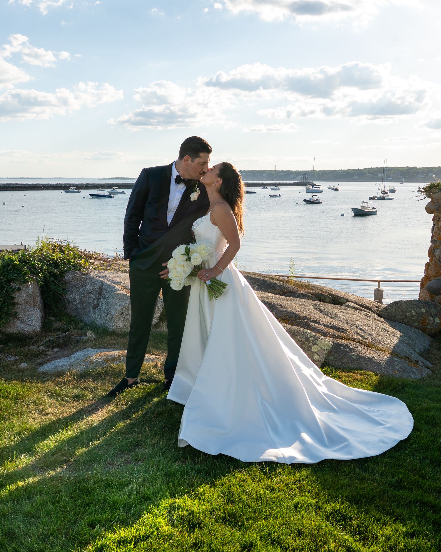 Bride and groom share a romantic kiss by a scenic coastal waterway, she holds white bouquet in elegant satin gown.
