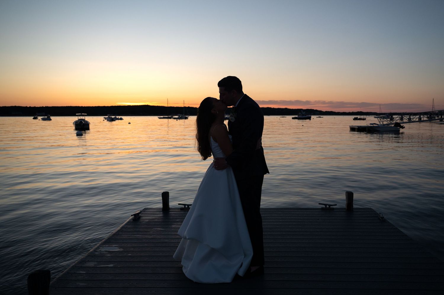 Silhouette of bride and groom embracing on a dock at sunset with calm water and boats in the background.