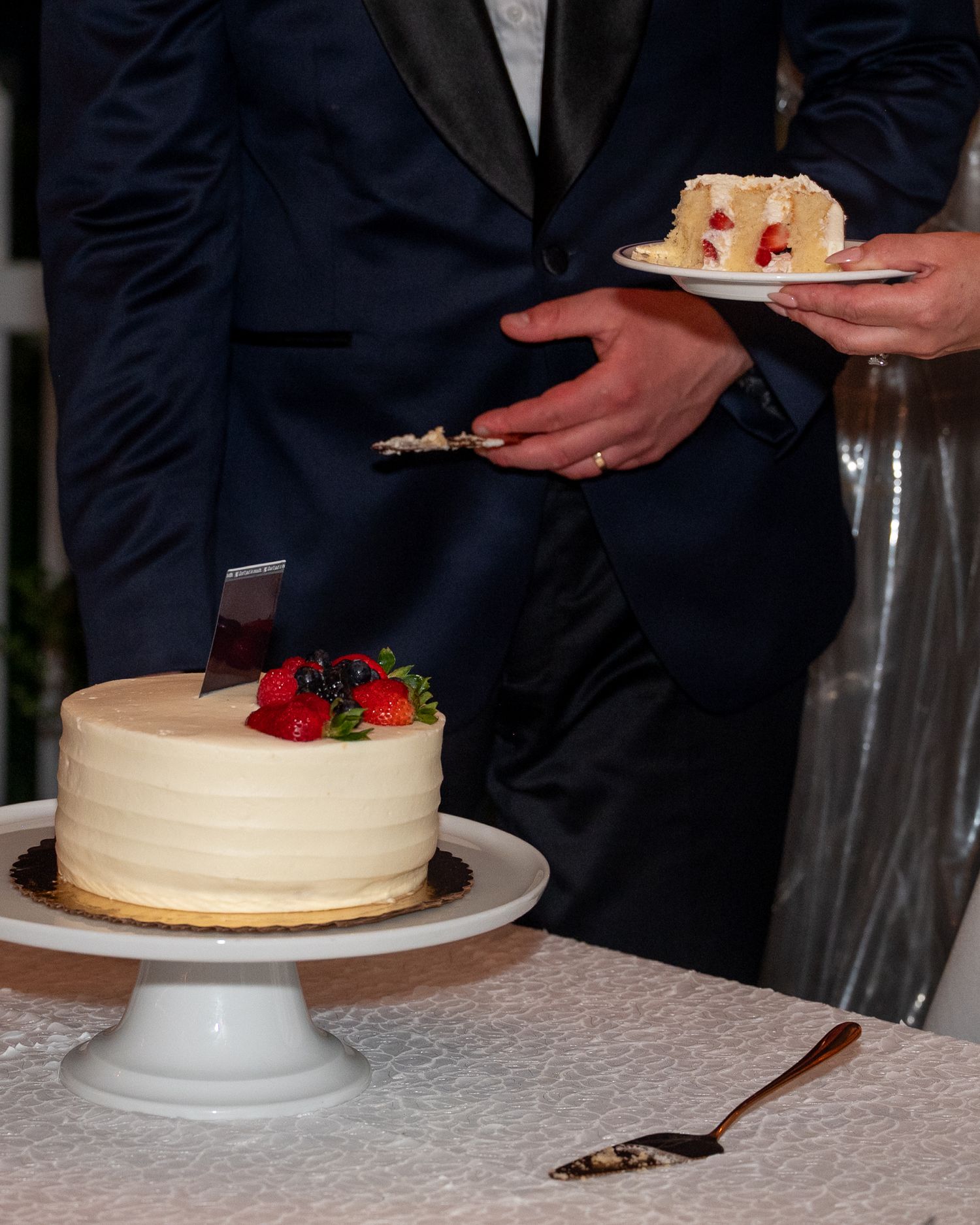 Two people in navy suits cutting a white frosted wedding cake topped with fresh strawberries and berries on a white pedestal.