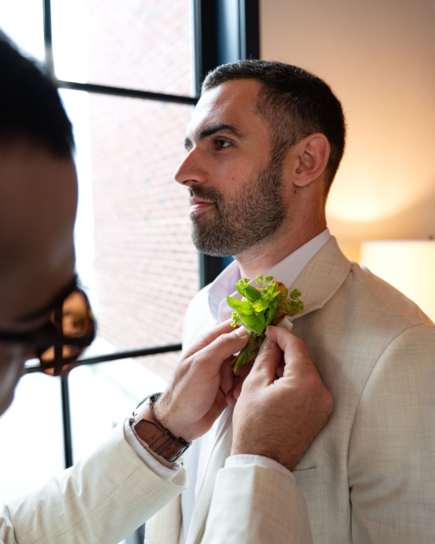 Someone has their boutonniere adjusted on their light colored suit jacket during what appears to be a wedding preparation.