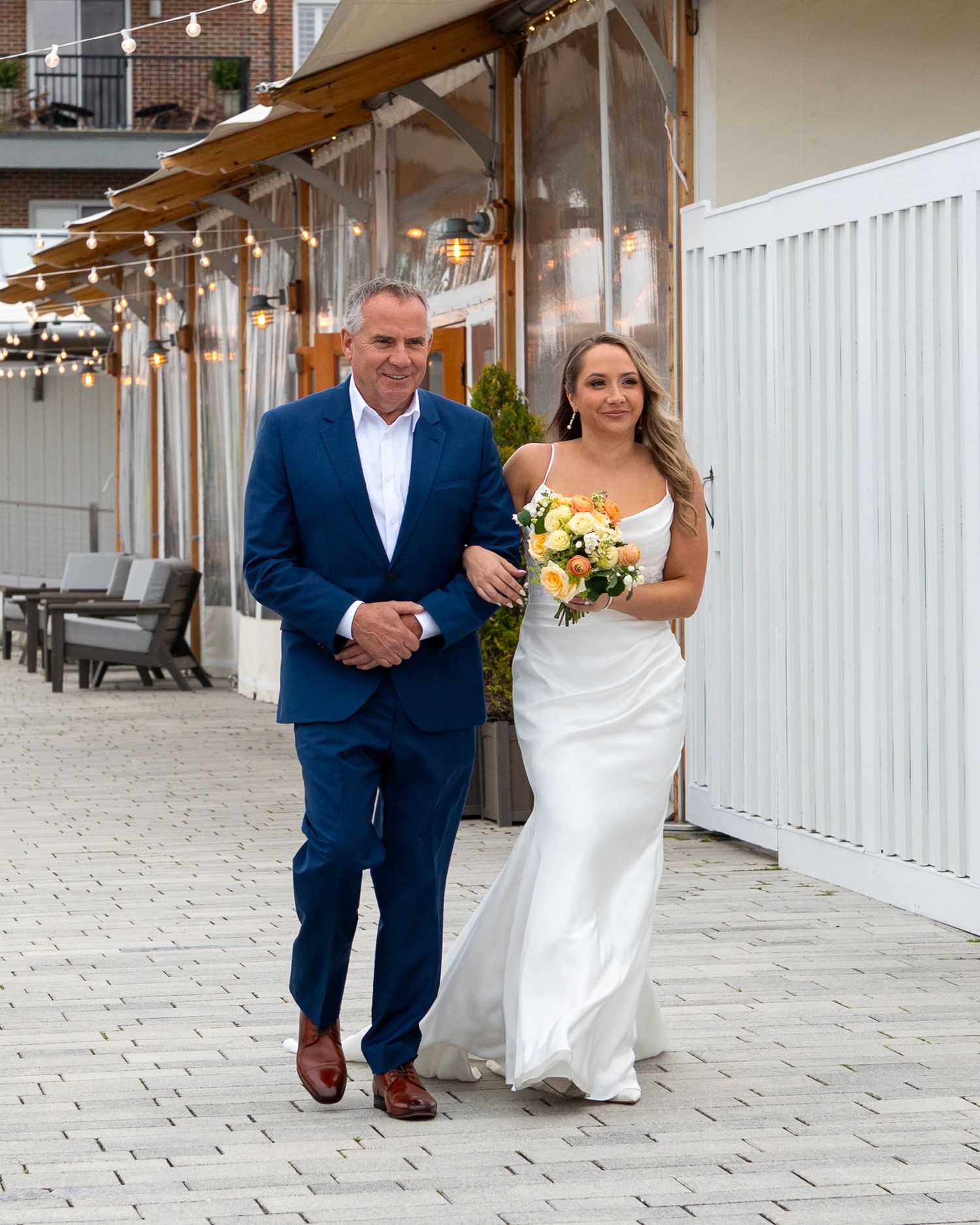 A happy couple walks arm in arm wearing a navy suit and white dress along a covered outdoor corridor.