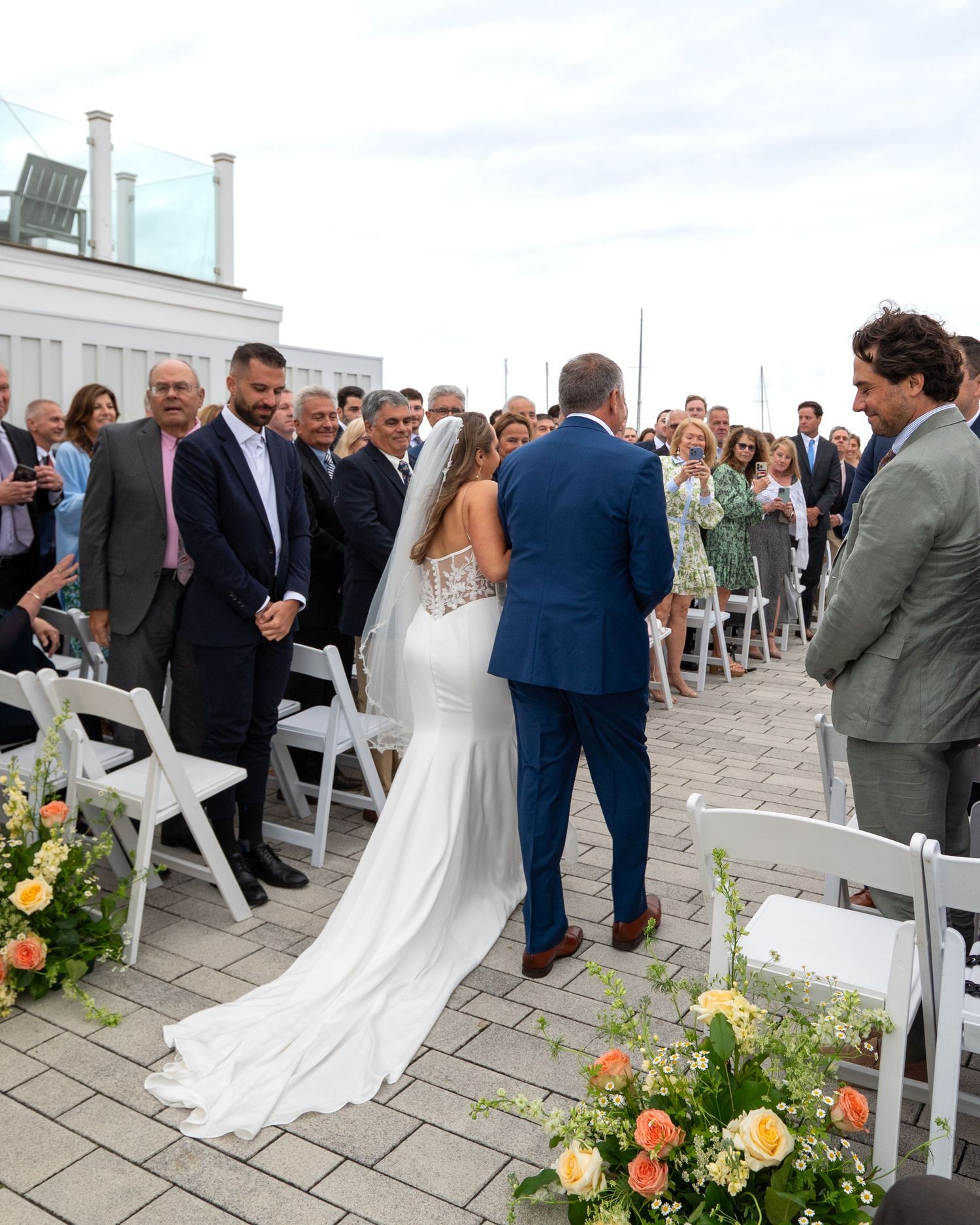 Wedding ceremony on a waterfront deck with white chairs and peach floral decorations lining the aisle.