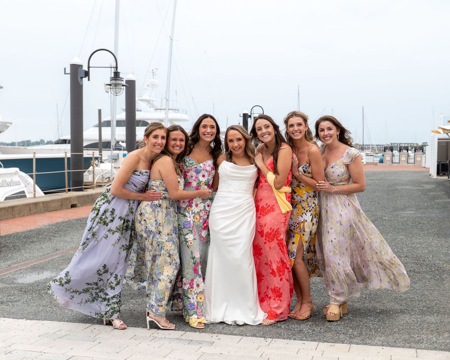 A group in colorful floral dresses pose together on a dock with boats and water in the background.