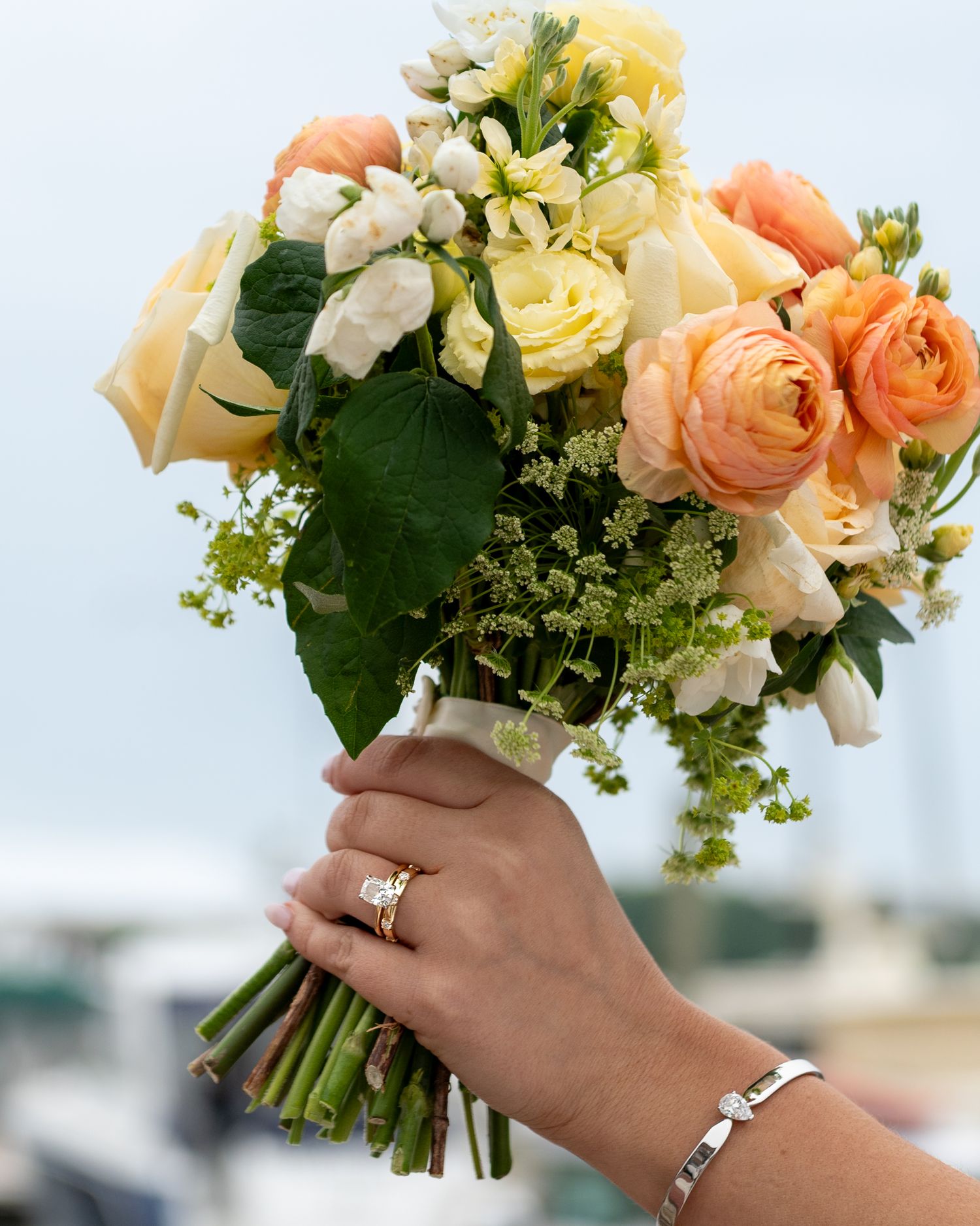 A delicate bridal bouquet of peach roses and white flowers with greenery, shown with jewelry-adorned hand holding stems.