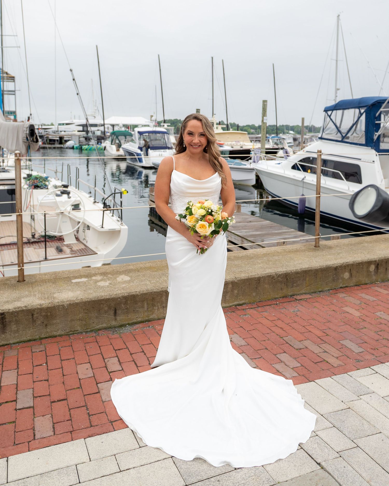 A bride in a flowing white wedding dress stands on a brick walkway near a marina with boats in the background.