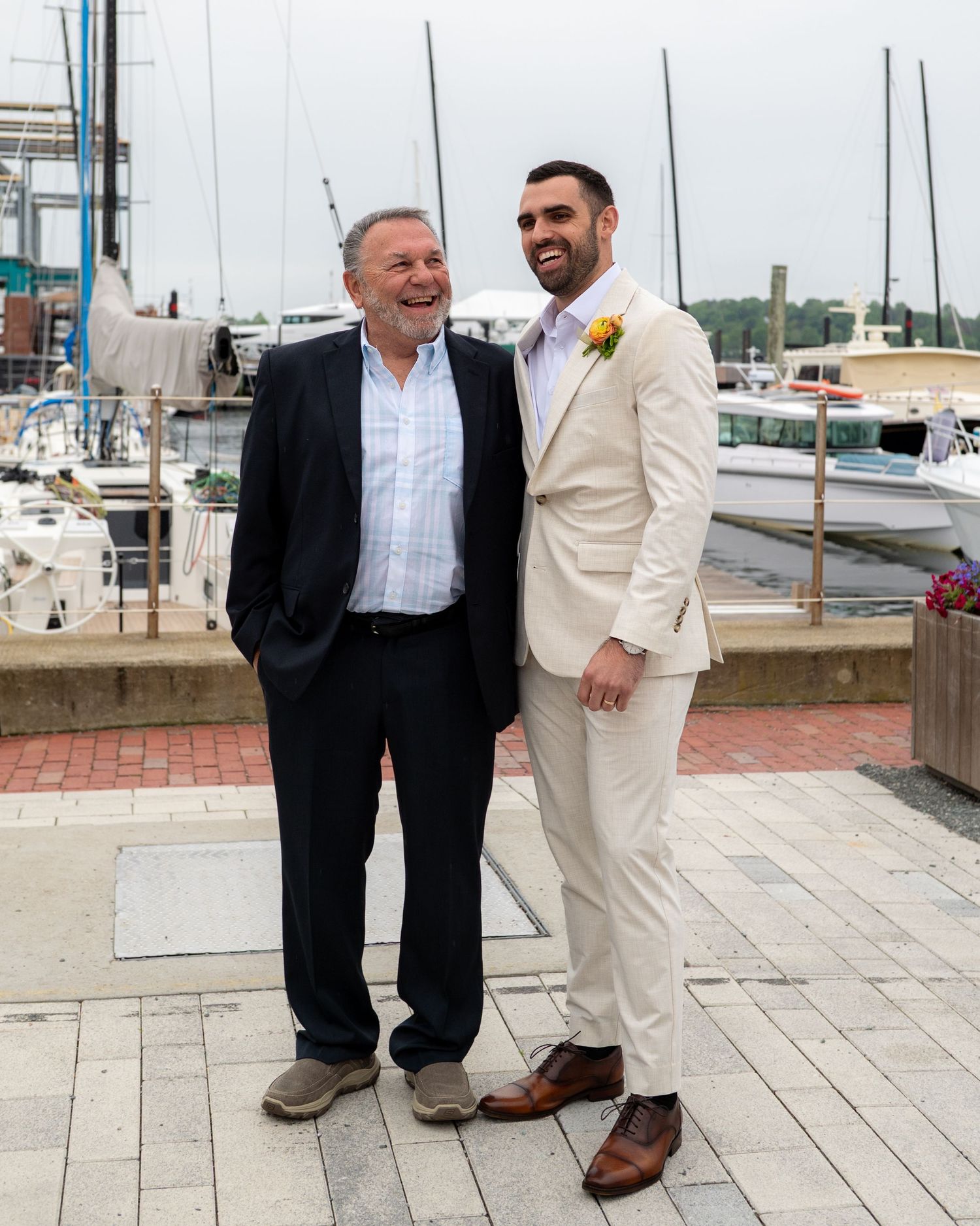 Two people in formal attire stand together smiling on a marina dock with boats and masts visible in the background.