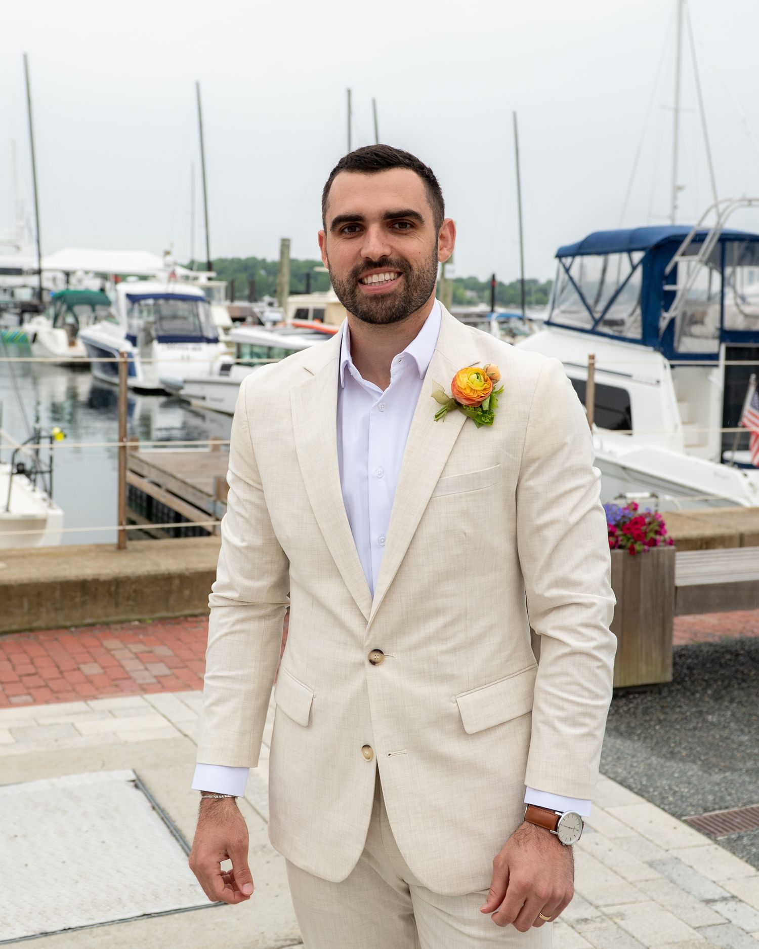 A groomsman in a beige suit with boutonniere stands by the waterfront with boats and a marina in the background.