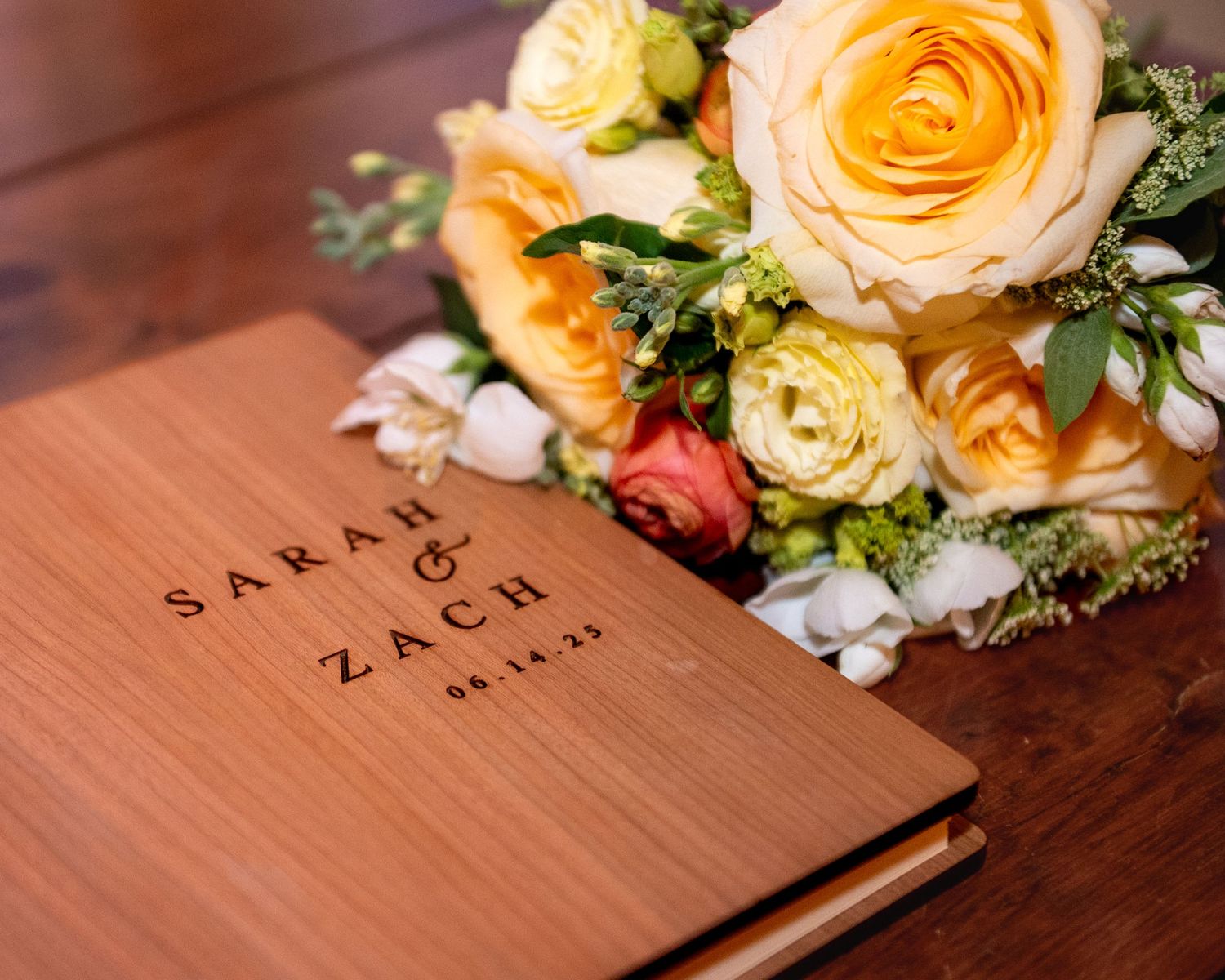 Peach and blush roses arranged in a elegant bouquet lay beside a wooden surface with engraved names.