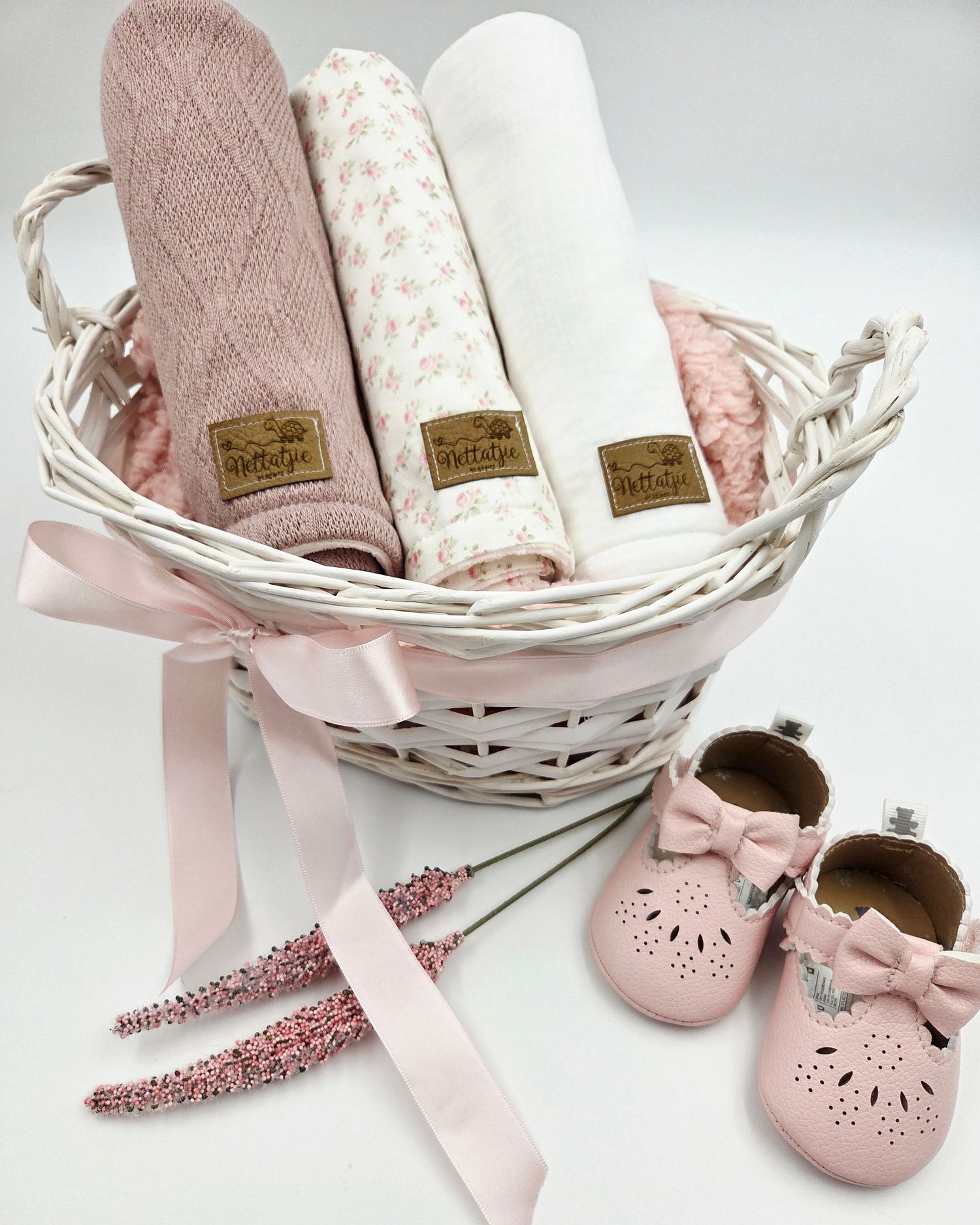 Pink baby blankets and shoes arranged in a white wicker basket with delicate ribbon accents.