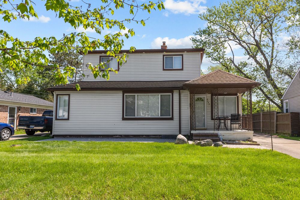 Two-story white sided home with brown roof, covered porch, lush green yard, and mature trees in background.