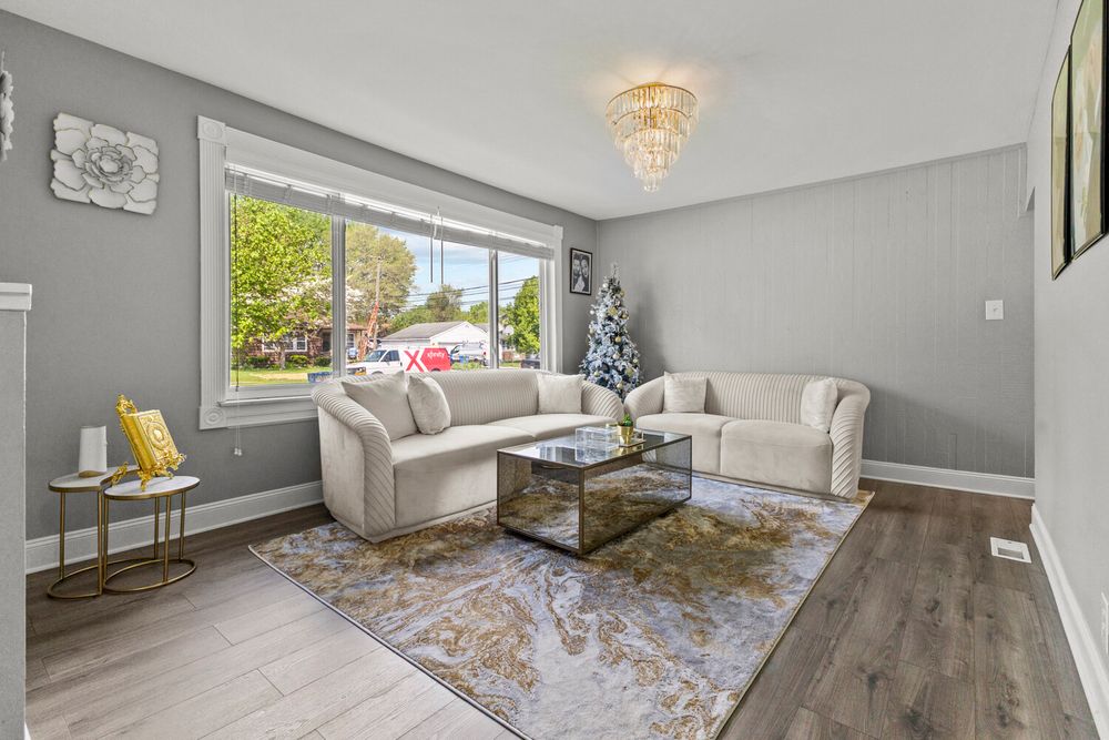Bright living room with gray walls, white sofas, glass coffee table, decorative rug, and large front window.