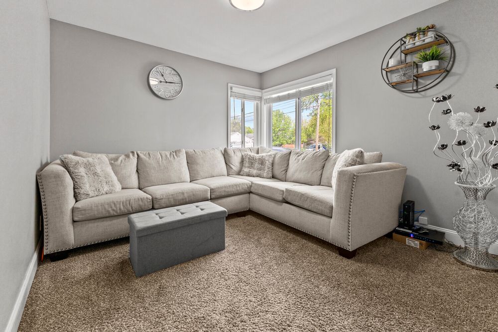 Cozy carpeted living room with large L-shaped gray sectional sofa, ottoman, and natural light from window.