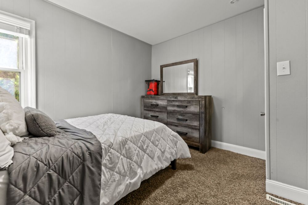 Simple bedroom featuring dark wood dresser with mirror, gray bedding, and carpet floors.