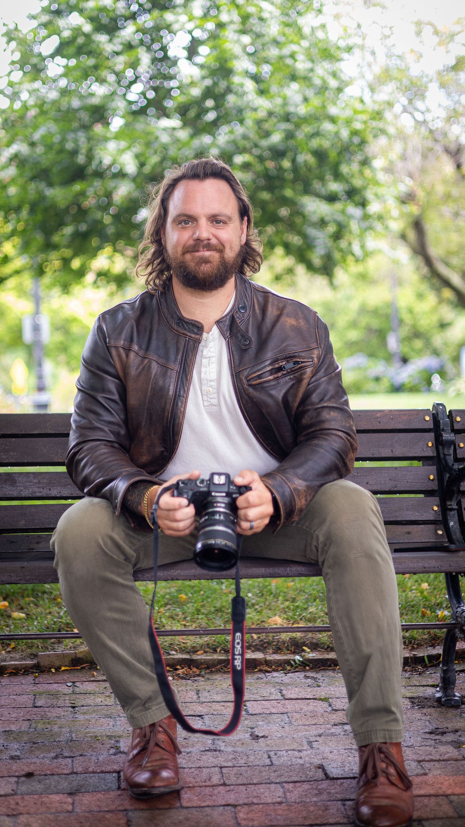 Photographer in leather jacket sitting on park bench holding professional camera.