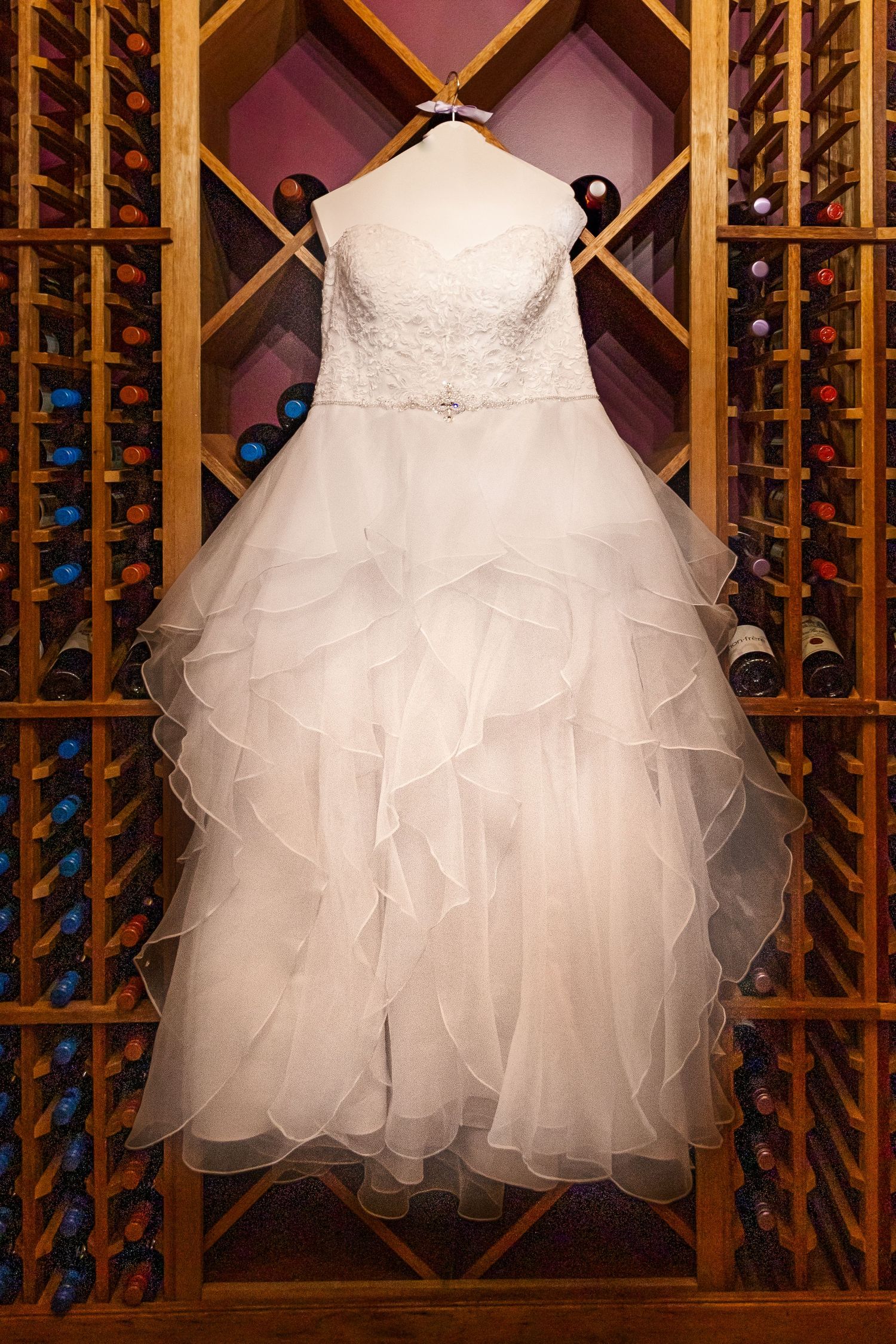 A stunning white wedding dress with lace bodice and ruffled tulle skirt displayed on wooden wine rack.