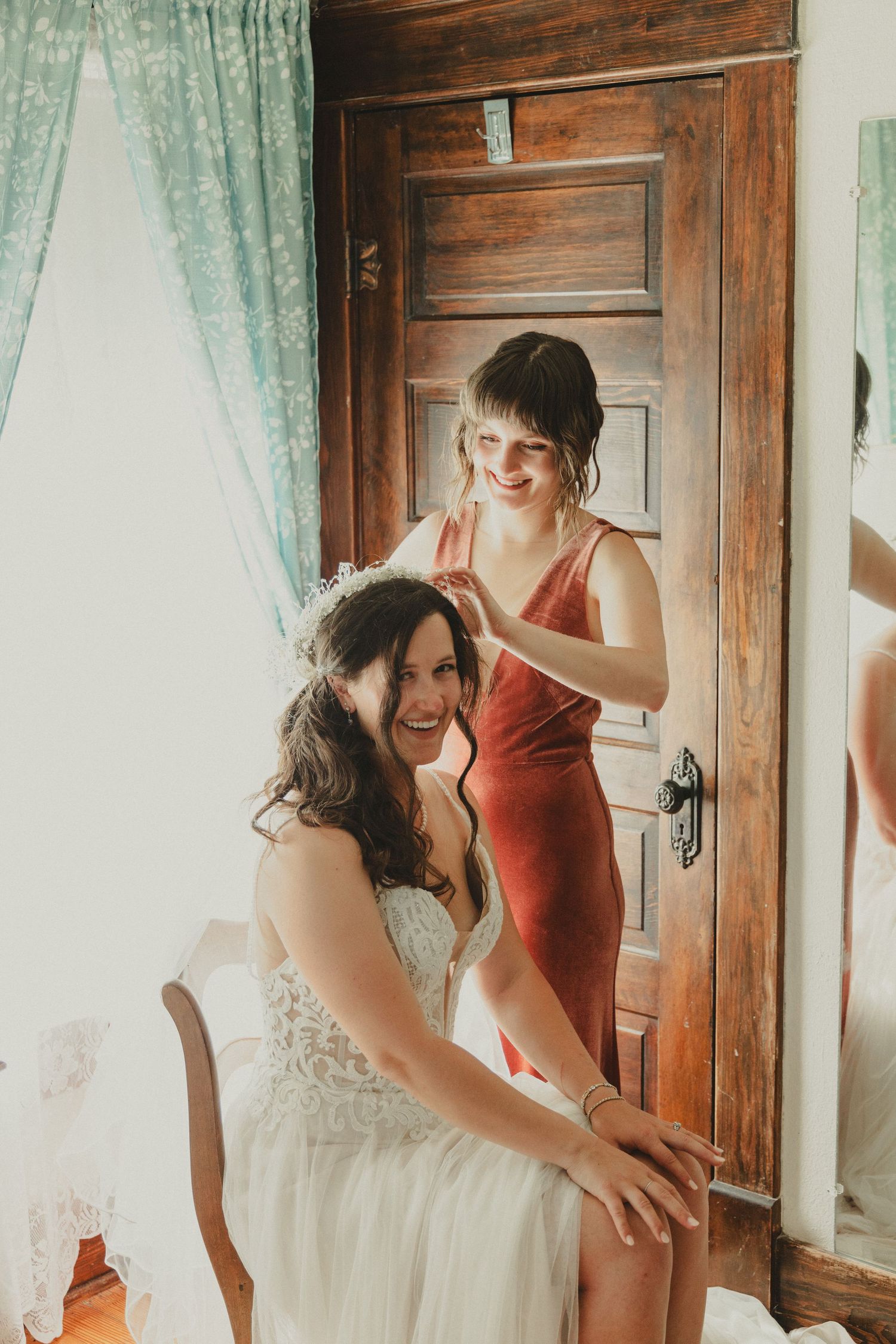 Getting ready moment between bride and attendant by wooden door with mint curtains.
