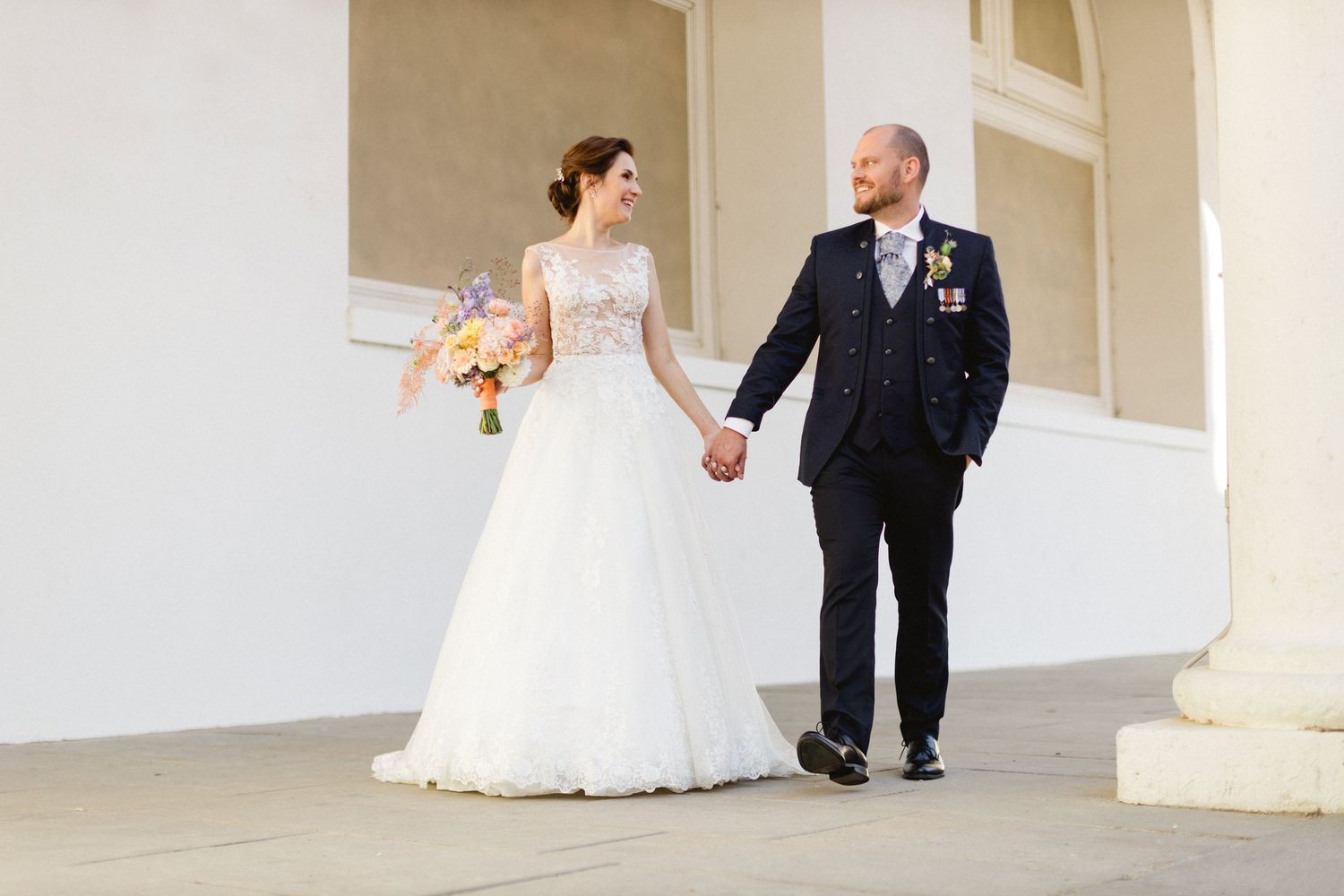 A bride wearing a lace white wedding dress walks hand in hand with a man in a black suit on a white pillared porch.