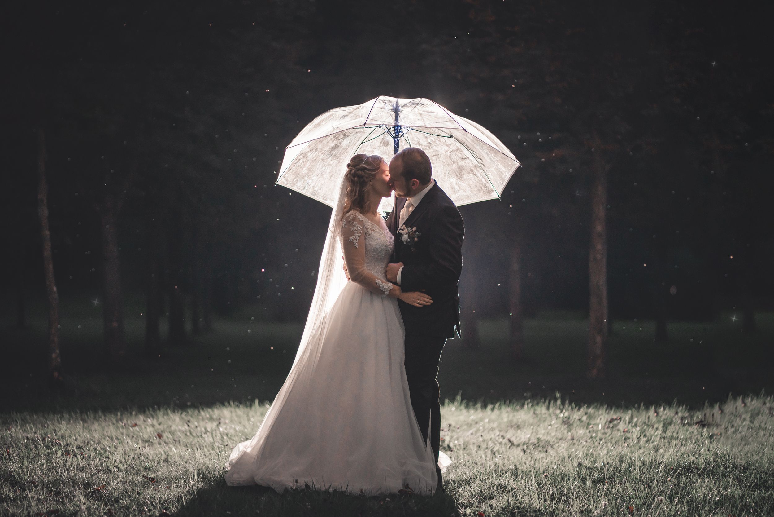 A wedding couple stands under an illuminated umbrella at night in a dreamy setting.