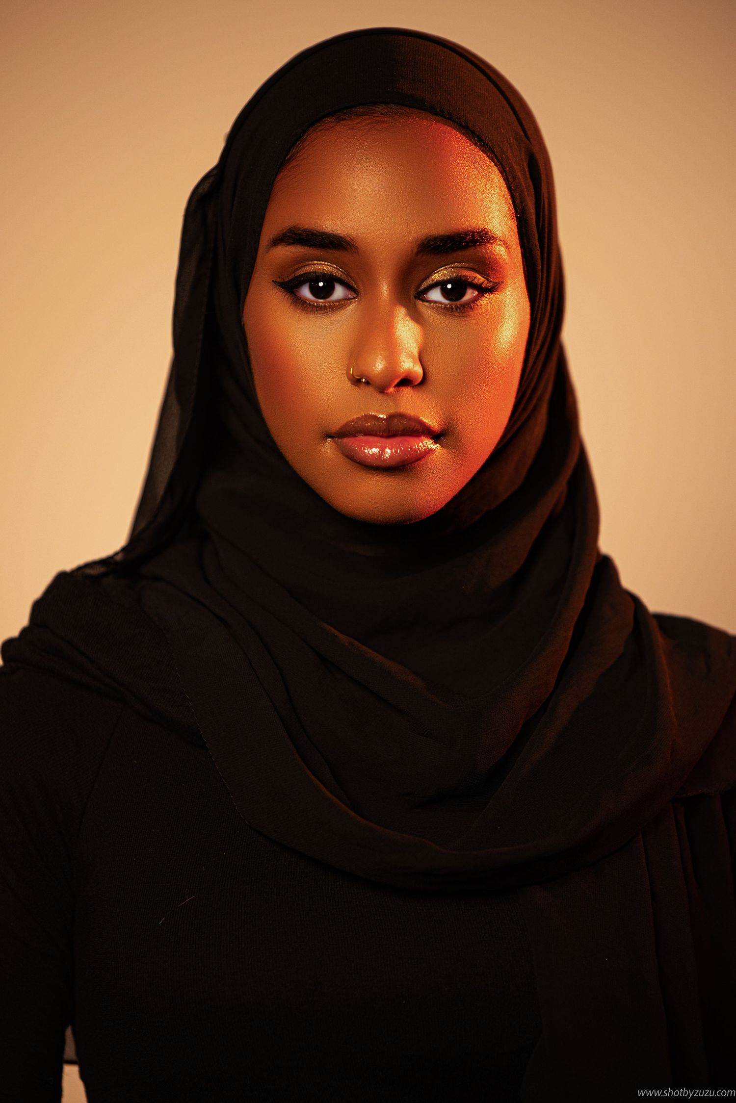 Portrait of a woman in black hijab with warm lighting and flawless makeup against bronze background.