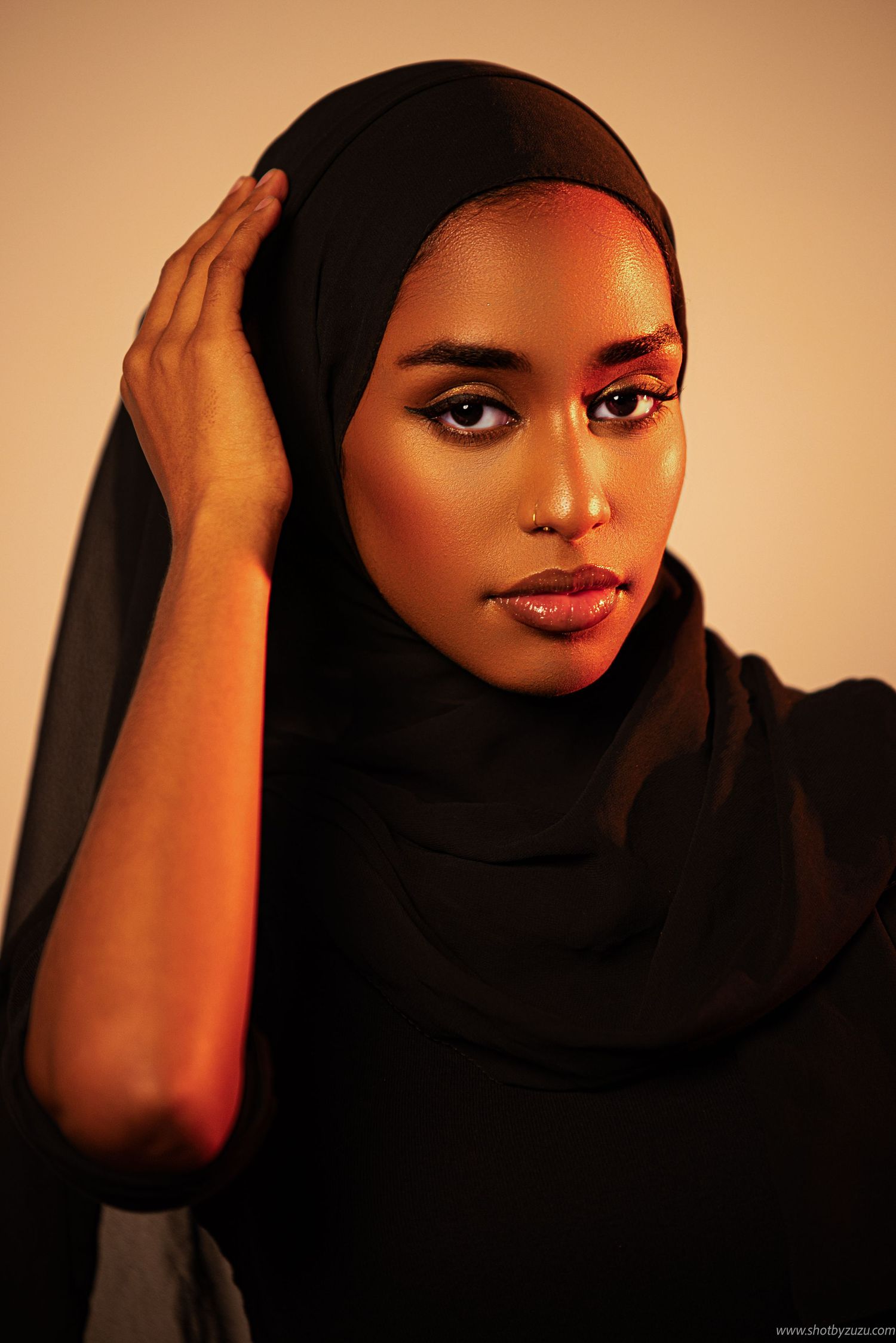 Artistic portrait of a woman in black hijab touching her head against warm-toned background.