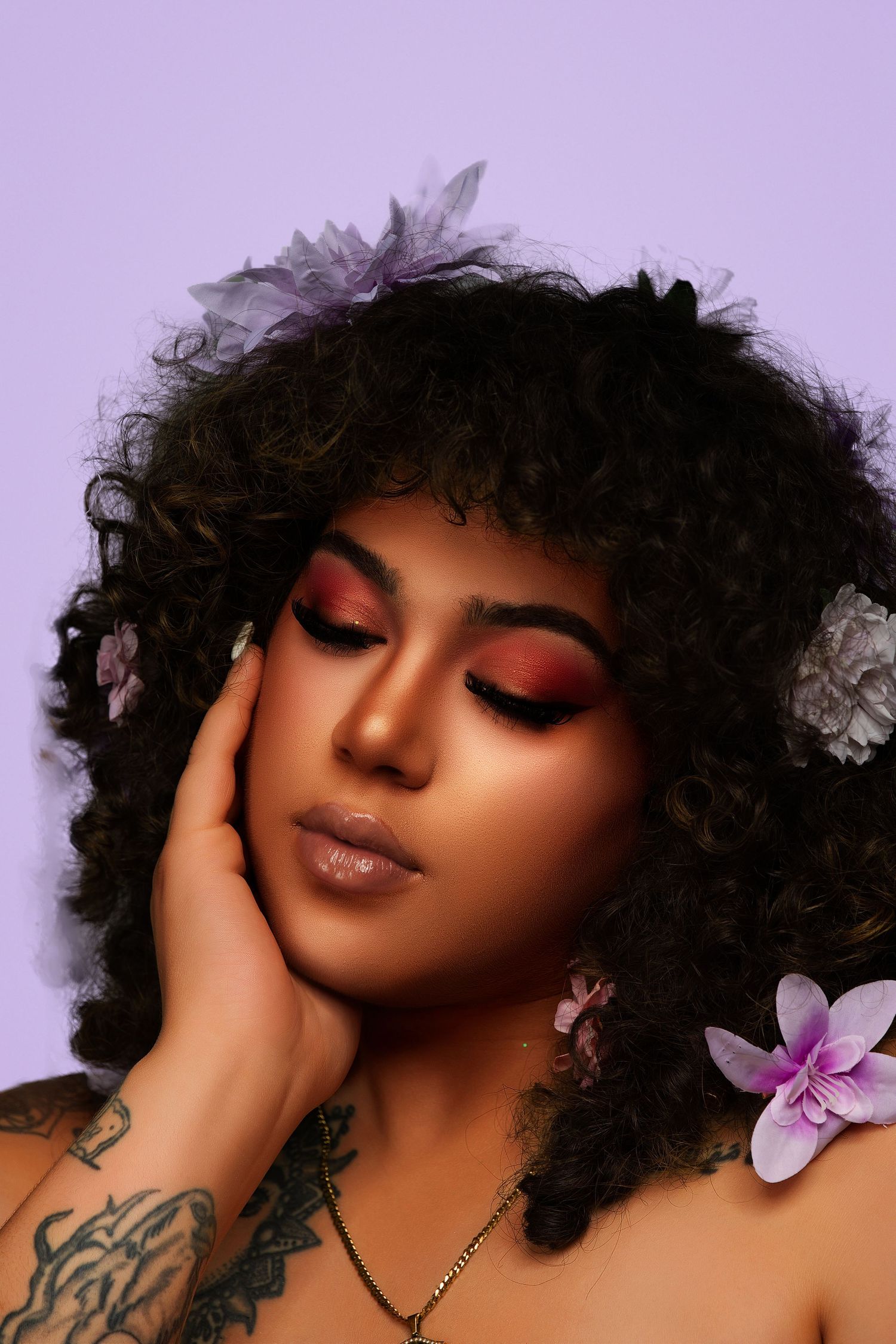 Beauty portrait with purple flowers, natural curls, and glowing makeup against lavender background.
