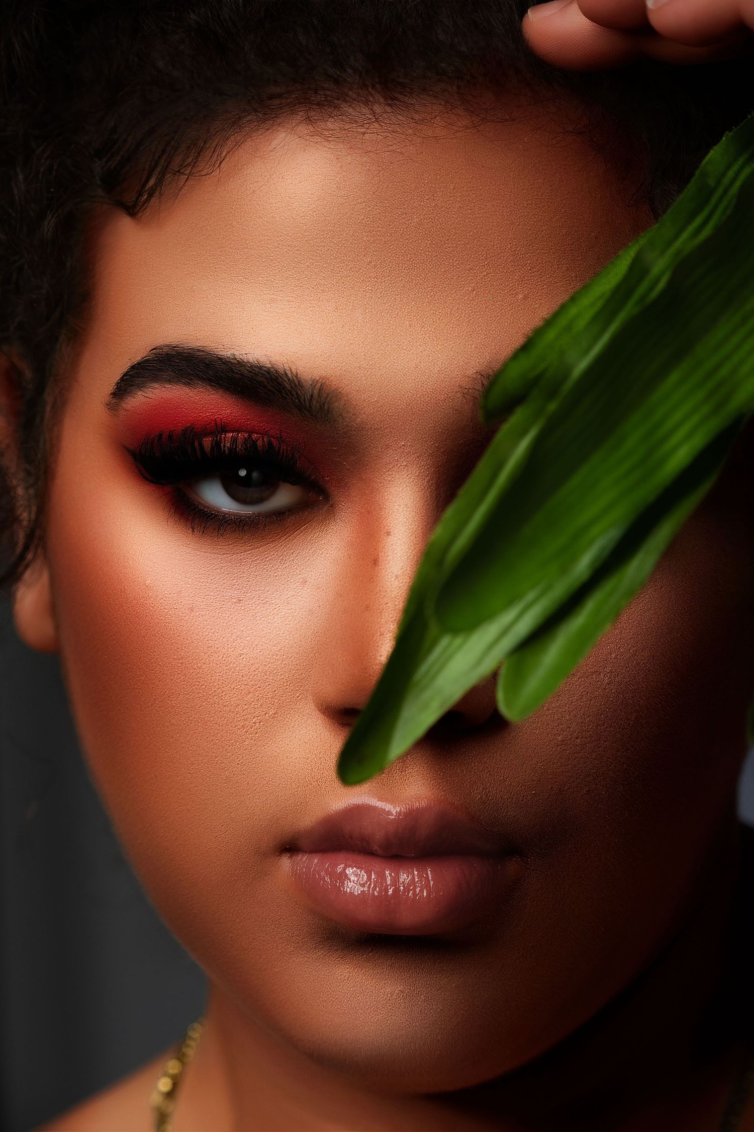 Creative beauty portrait with dramatic red eye makeup and green leaf accent against dark background.