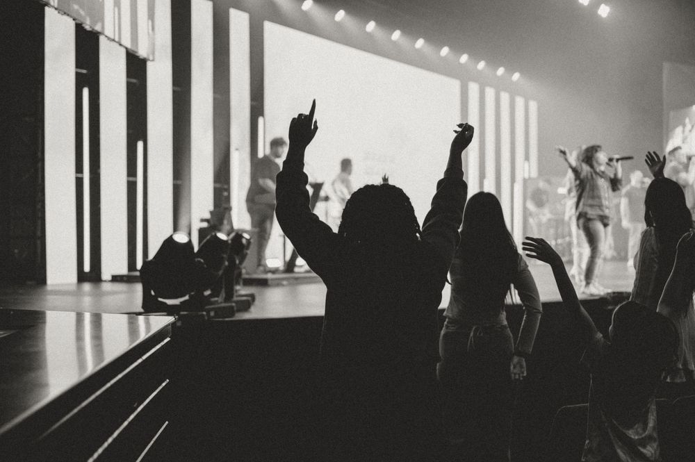 Silhouettes of concert attendees with raised hands against bright white stage lighting and vertical panels.
