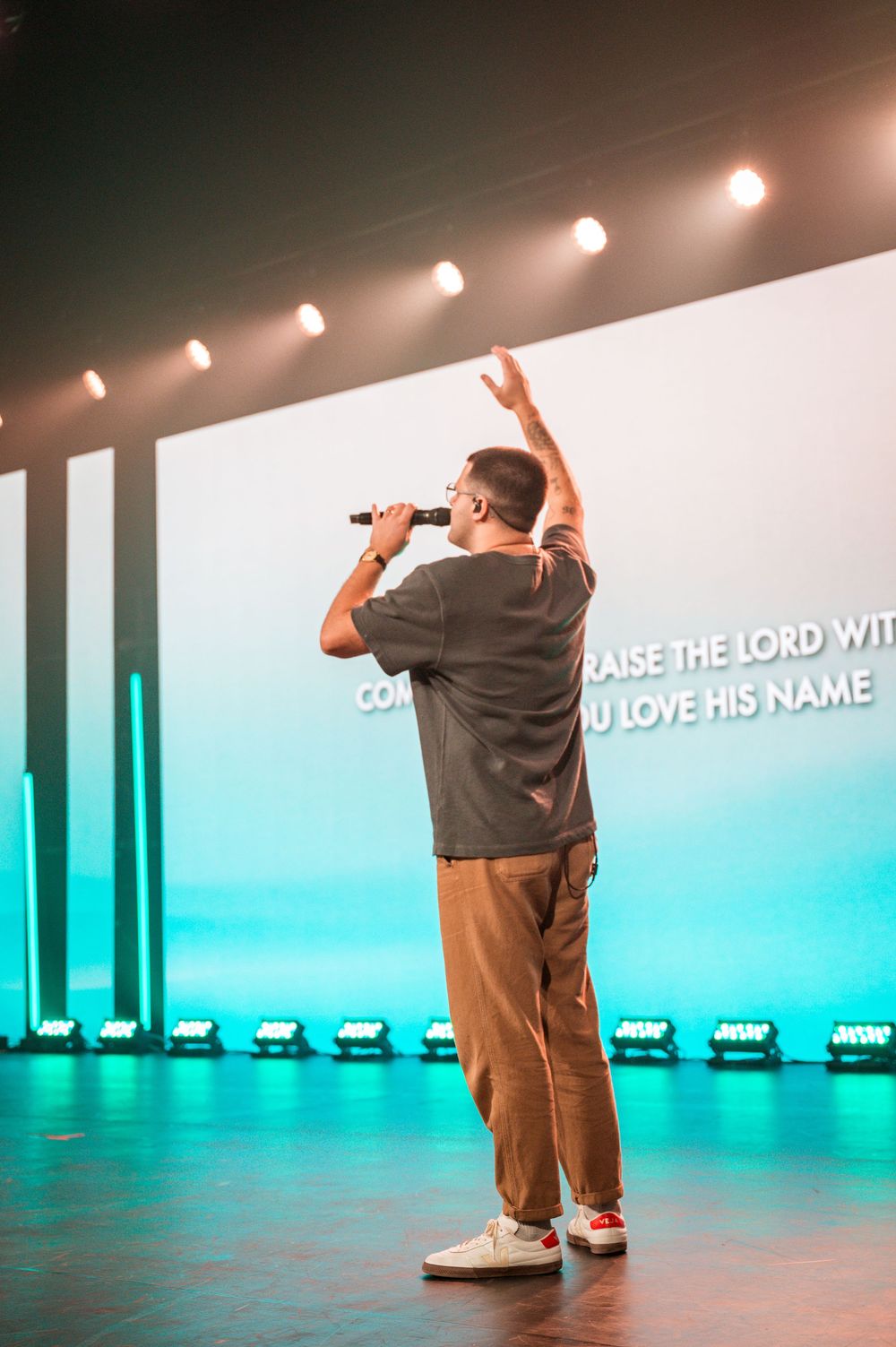 A person performs on stage with dramatic lighting and projected text about praising the Lord in the background.