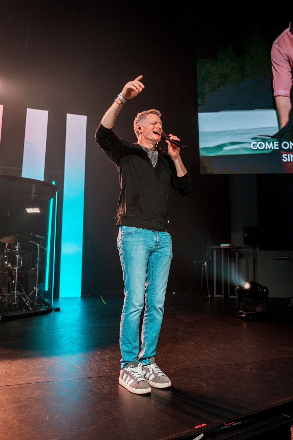 A performer in jeans and black sweater stands on stage with one arm raised, singing into a microphone under colorful lighting.