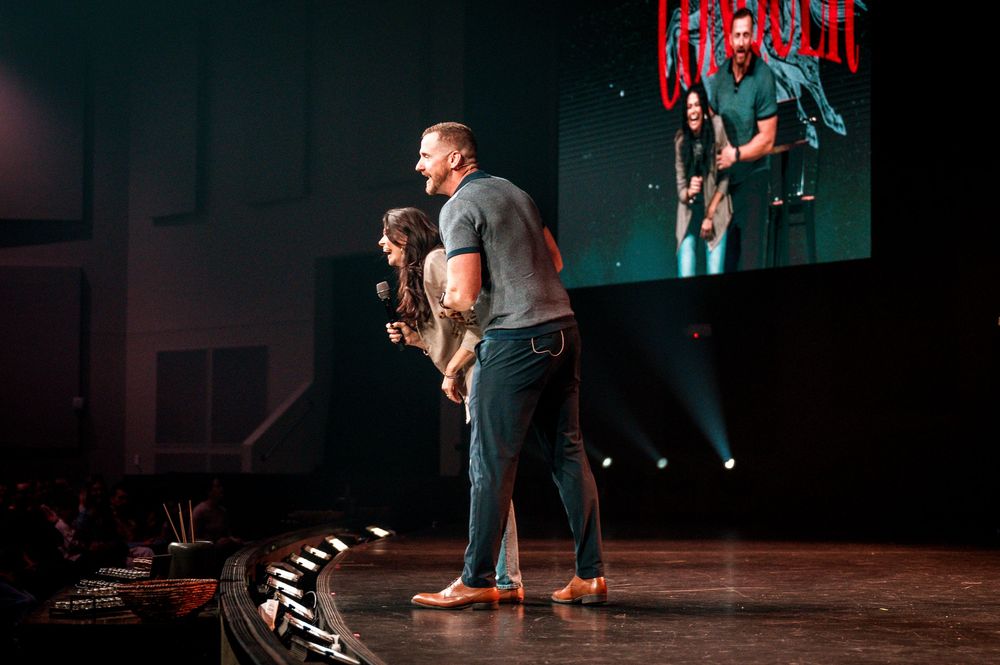 Performers on stage at a concert venue with dramatic lighting and video screens in the background.