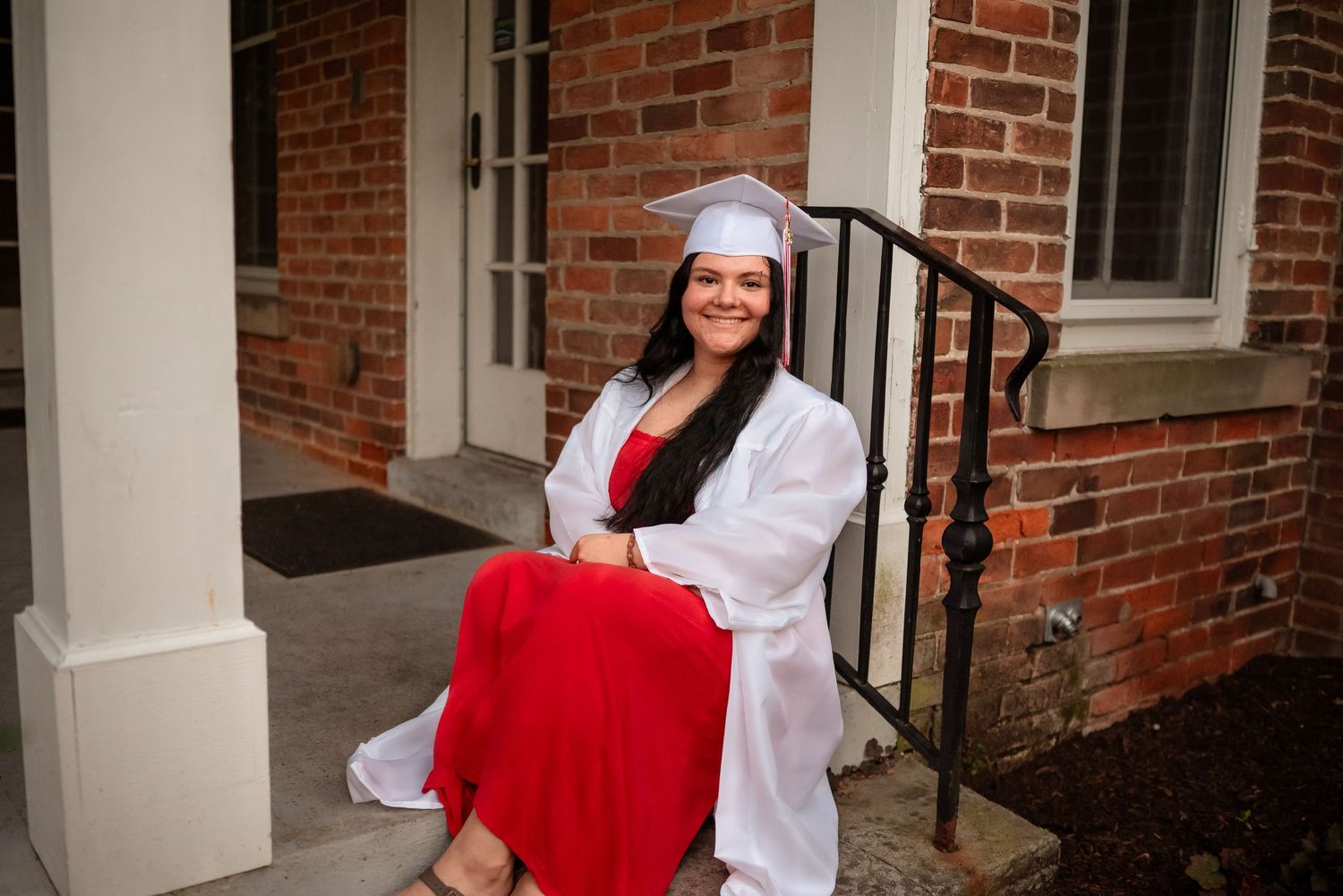 Graduate in red dress and white gown poses on brick steps of academic building.