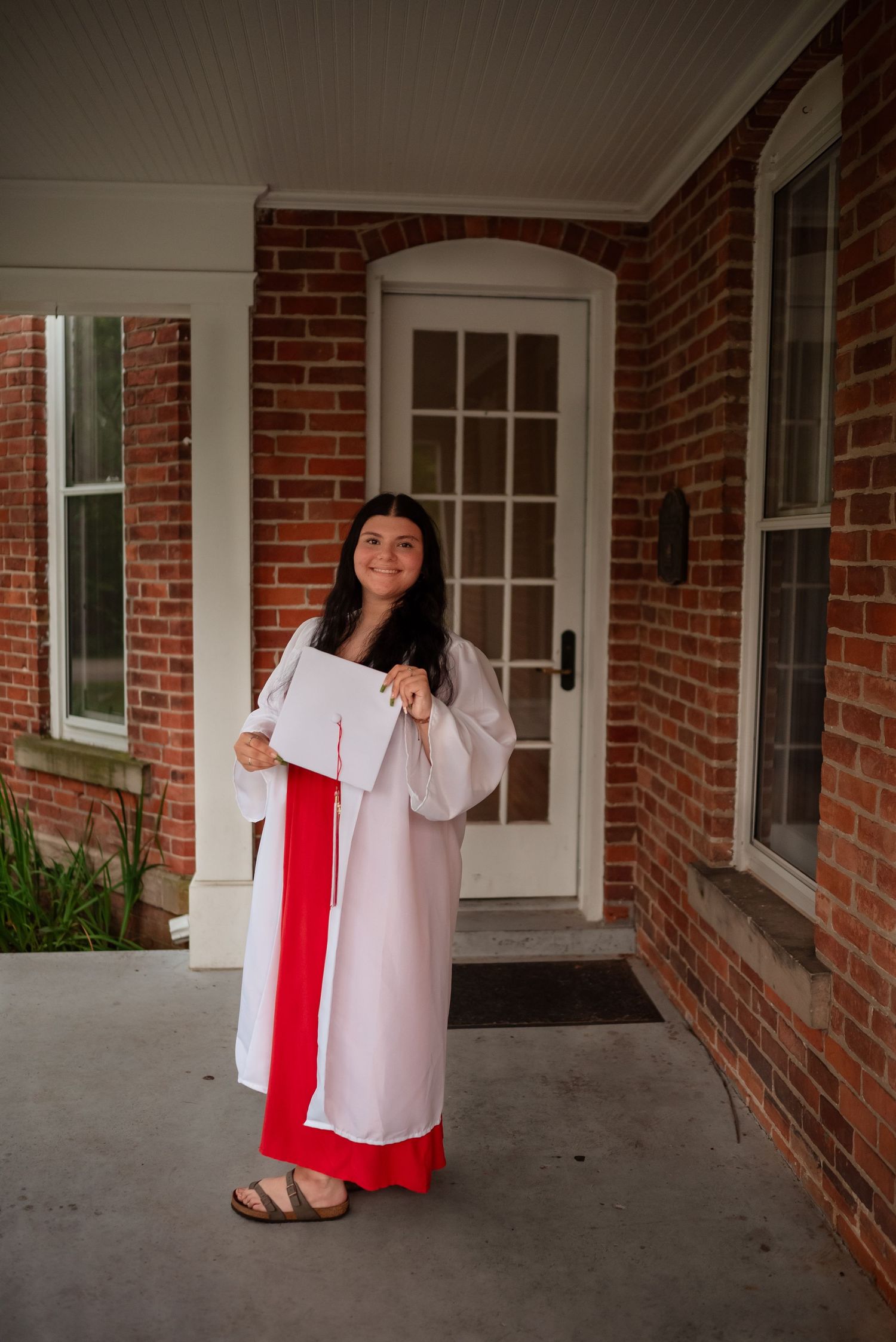 A graduate in white and red regalia poses playfully in front of a brick building entrance.