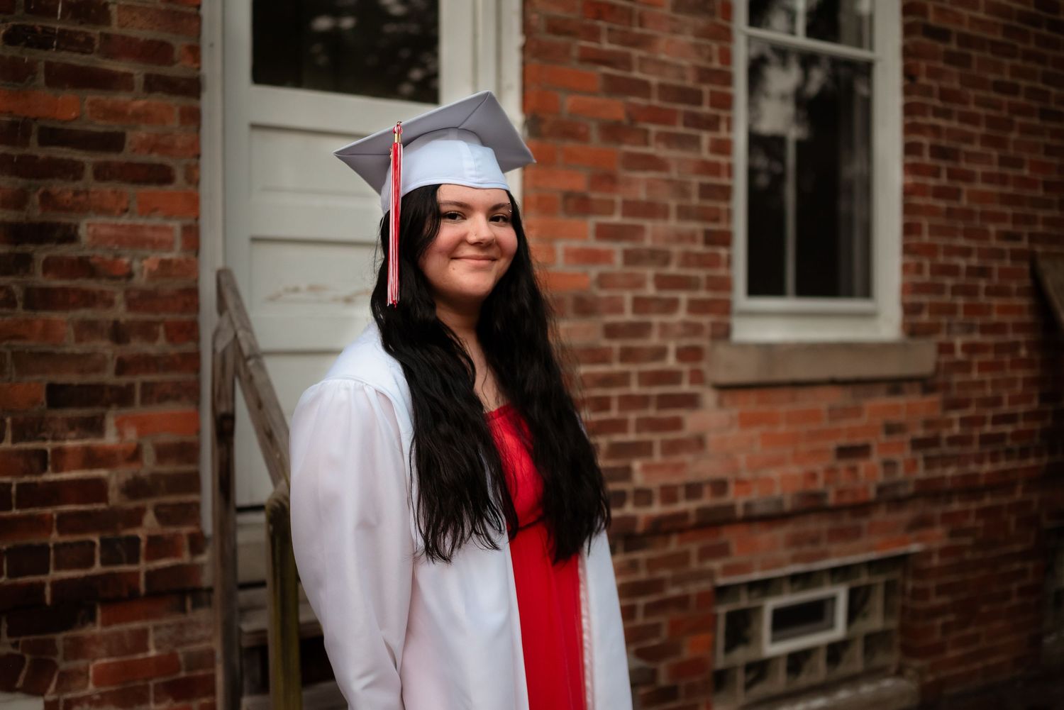 Graduate in white cap and gown with red stole stands smiling in front of brick building during graduation ceremony.