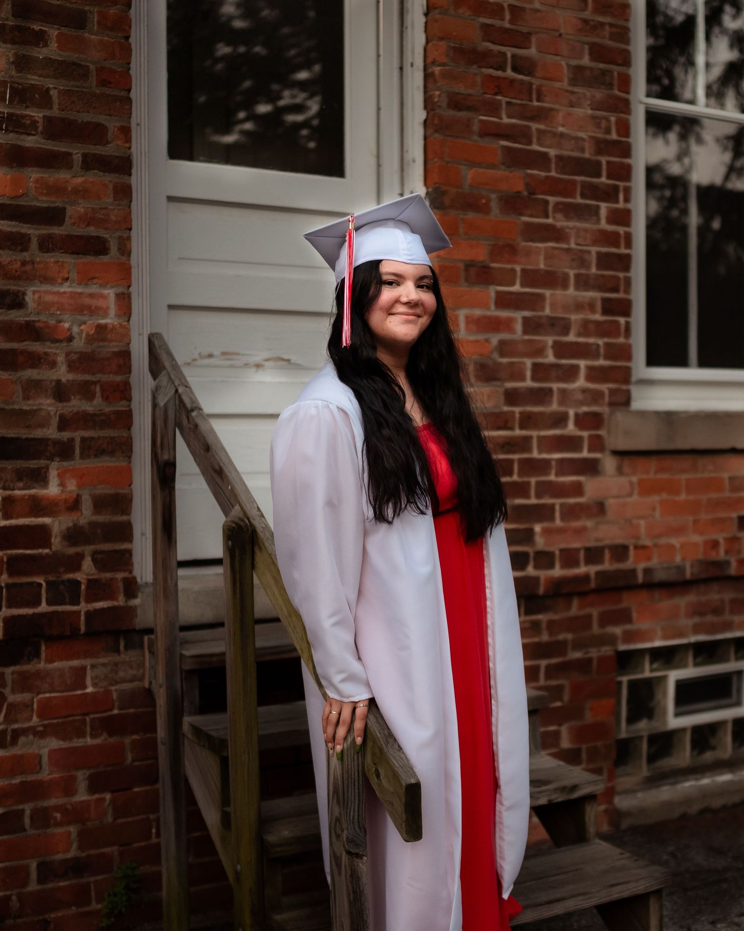 A graduate in white cap and gown with red stole poses against a red brick wall with white trim windows.