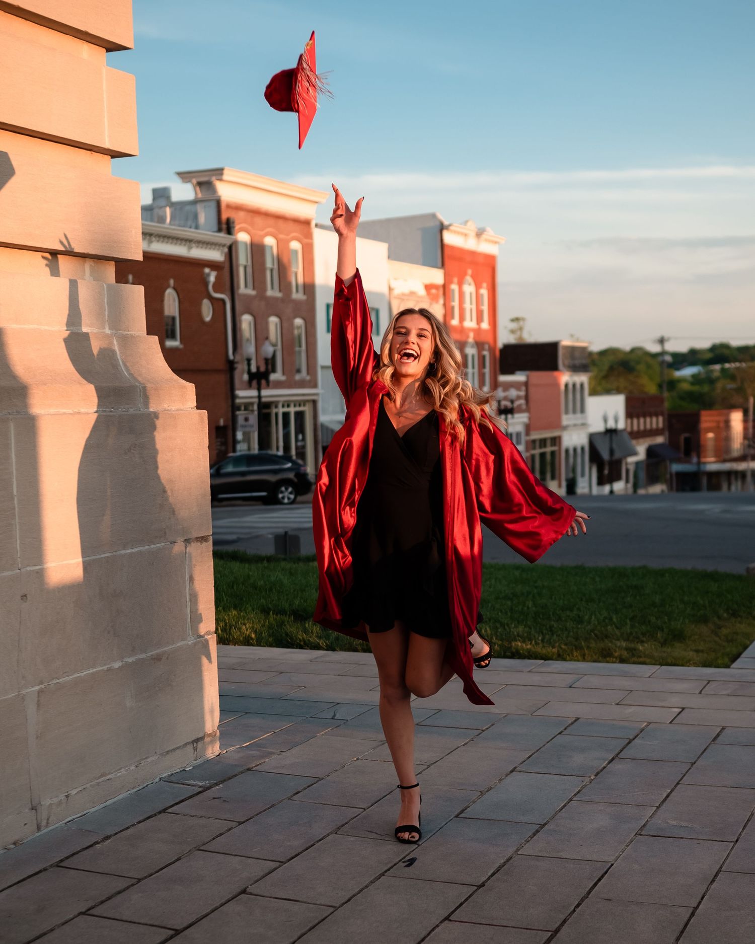 Graduate tosses red cap in air while celebrating on city street during sunset.