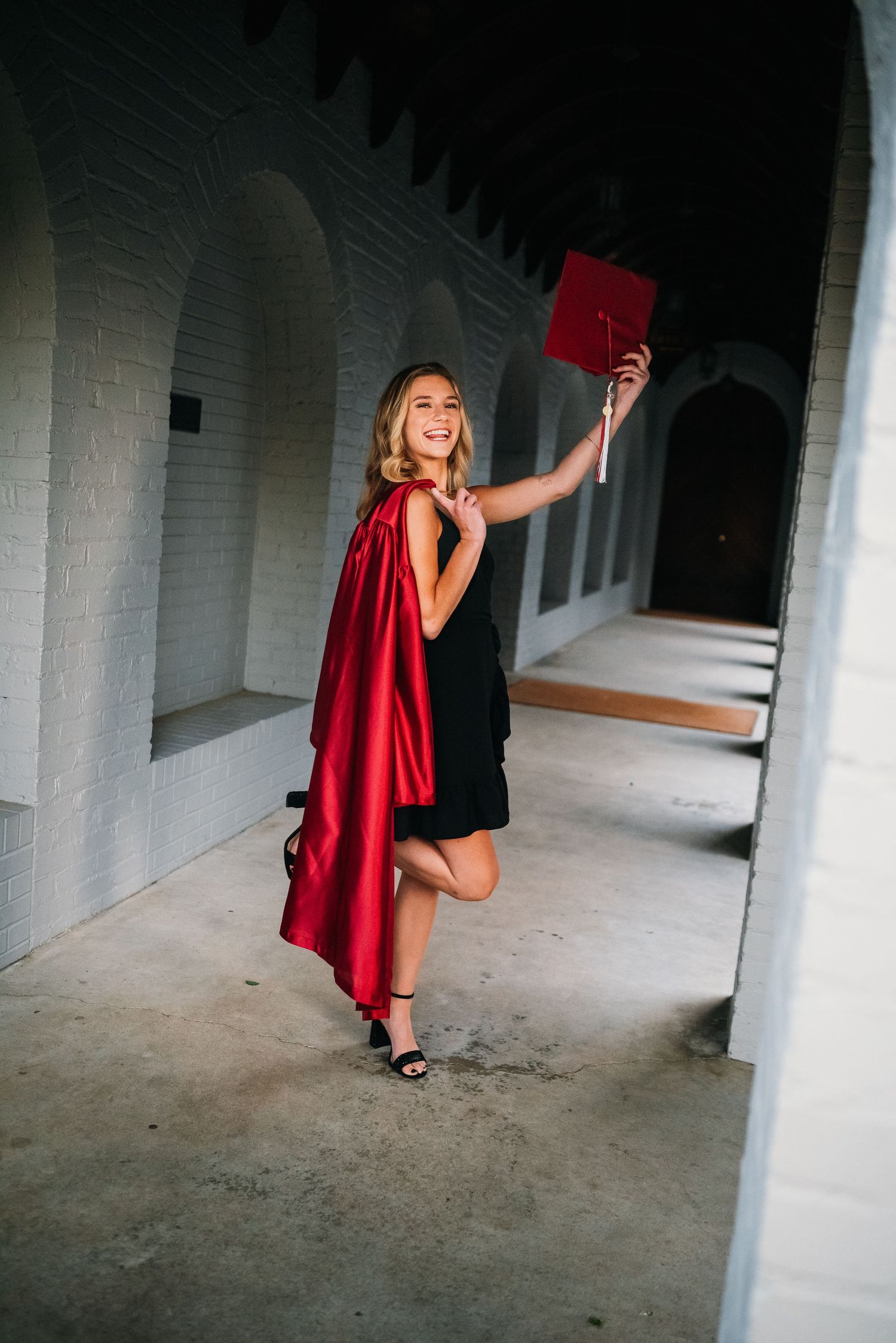 Graduate poses with diploma in arched hallway wearing red graduation gown.