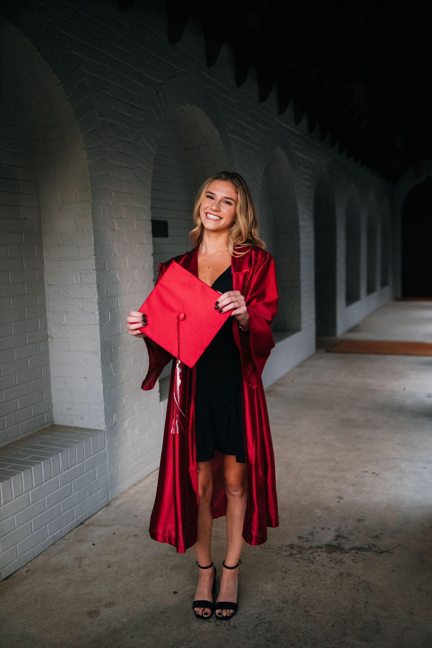 Graduate shows off diploma holder in hallway while wearing black dress and red graduation gown.