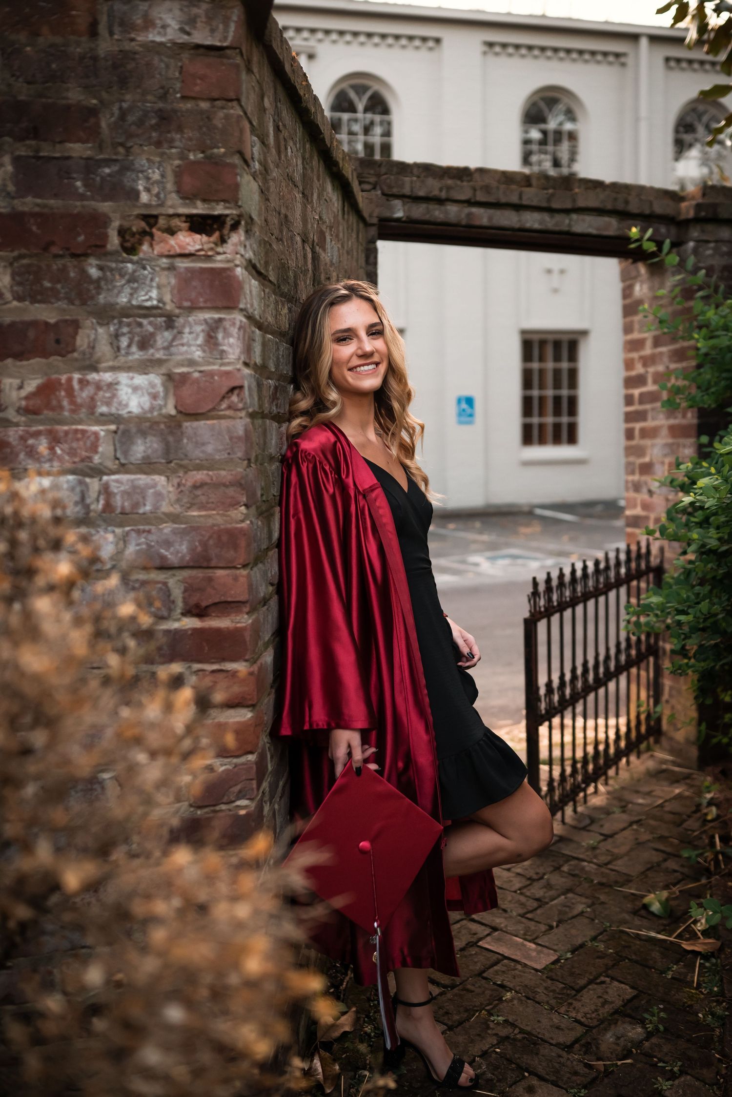 Graduate in red graduation attire poses against historic brick archway and iron fence.